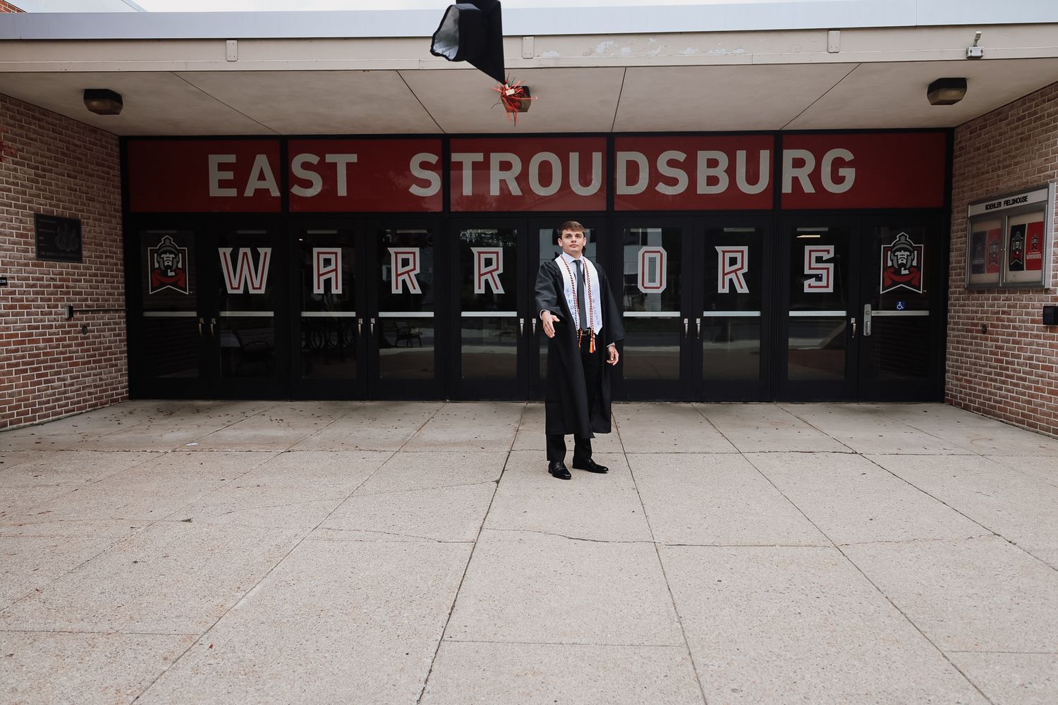 Graduate in cap and gown poses in front of East Stroudsburg Warriors school entrance on graduation day.