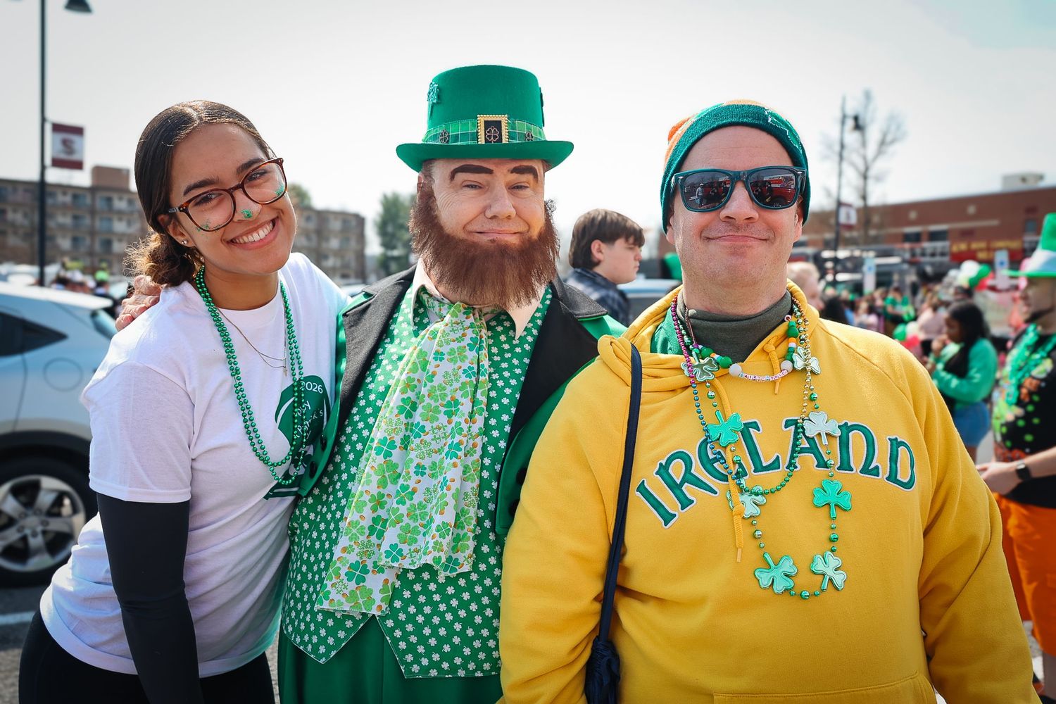 Three friends dressed in festive green St. Patrick's Day outfits smile together outdoors at a parade celebration.