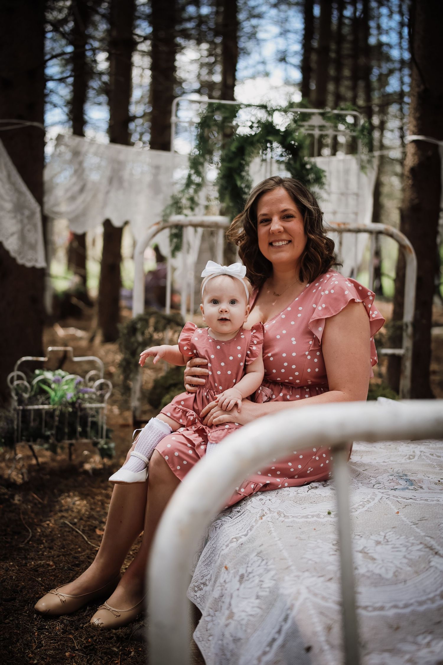 A woman and baby girl in matching pink polka dot dresses sit on a white bed frame in a woodland setting.