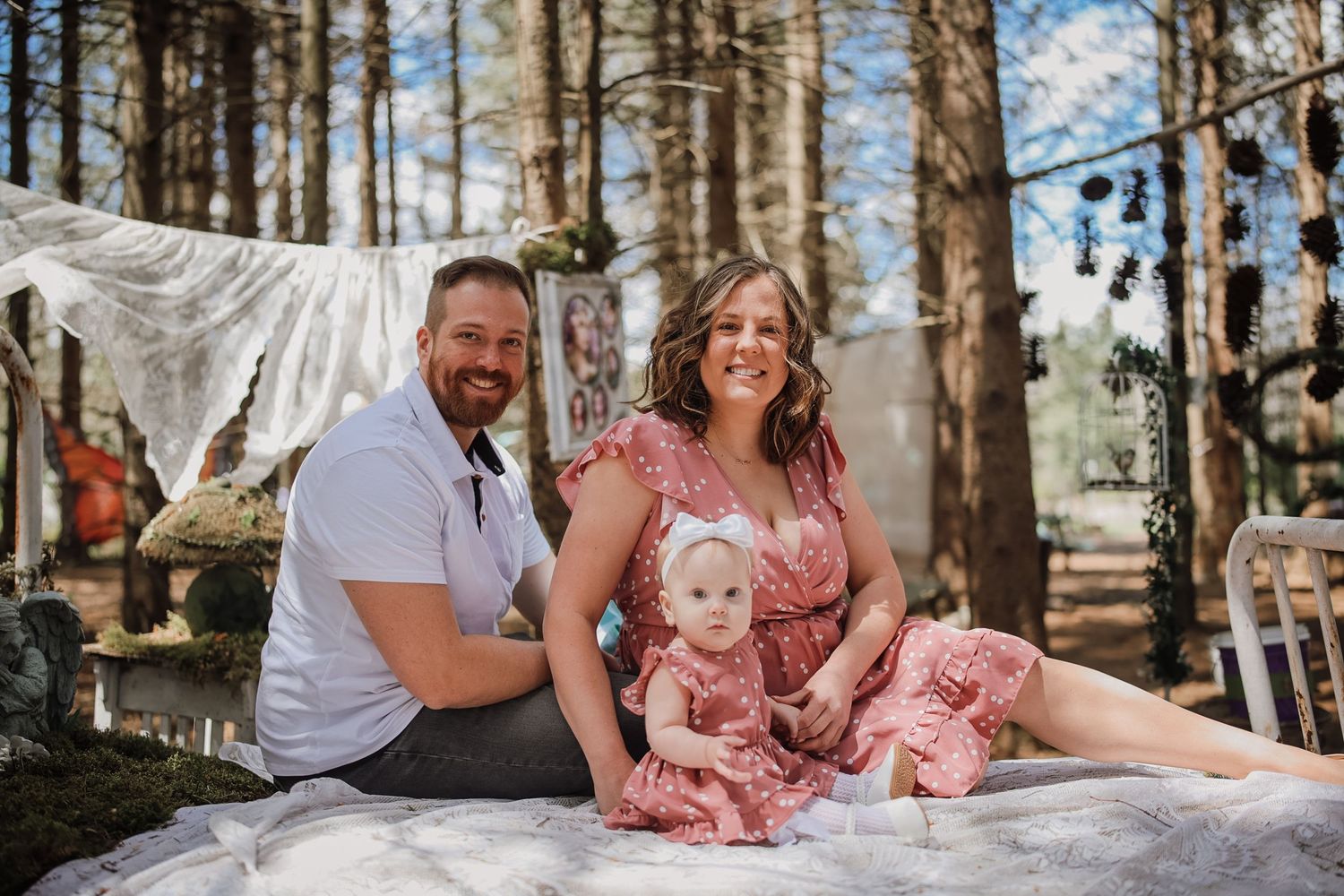 A smiling family of three poses outdoors in a wooded setting, with parents sitting beside their baby girl in a pink dress.