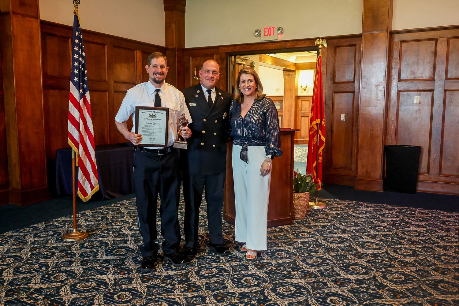 Three people posing with a certificate at a formal awards ceremony in an elegant wood-paneled room with American flag.