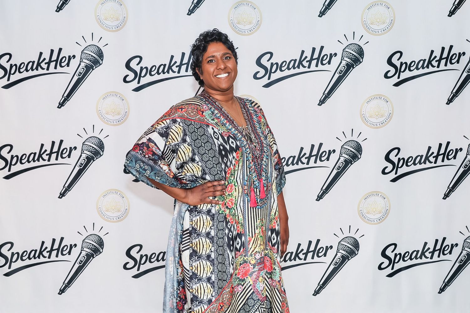 Woman in colorful patterned dress posing at SpeakHer event backdrop with microphone logo.
