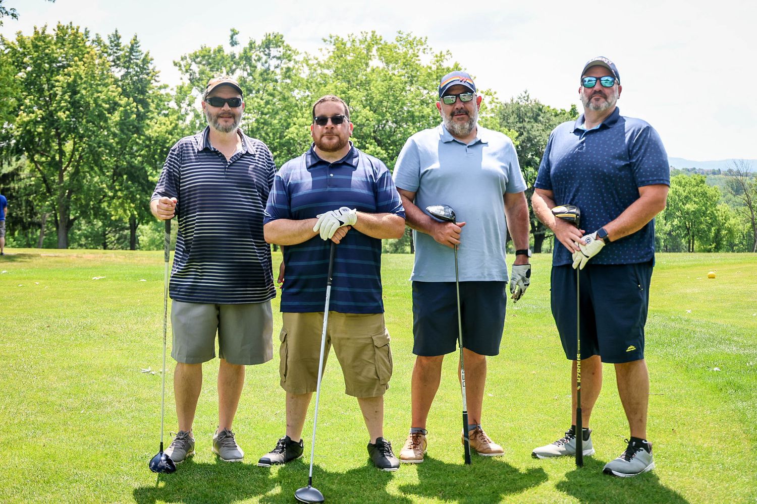 Four men posing together on a sunny golf course, holding clubs and smiling during a golf outing.