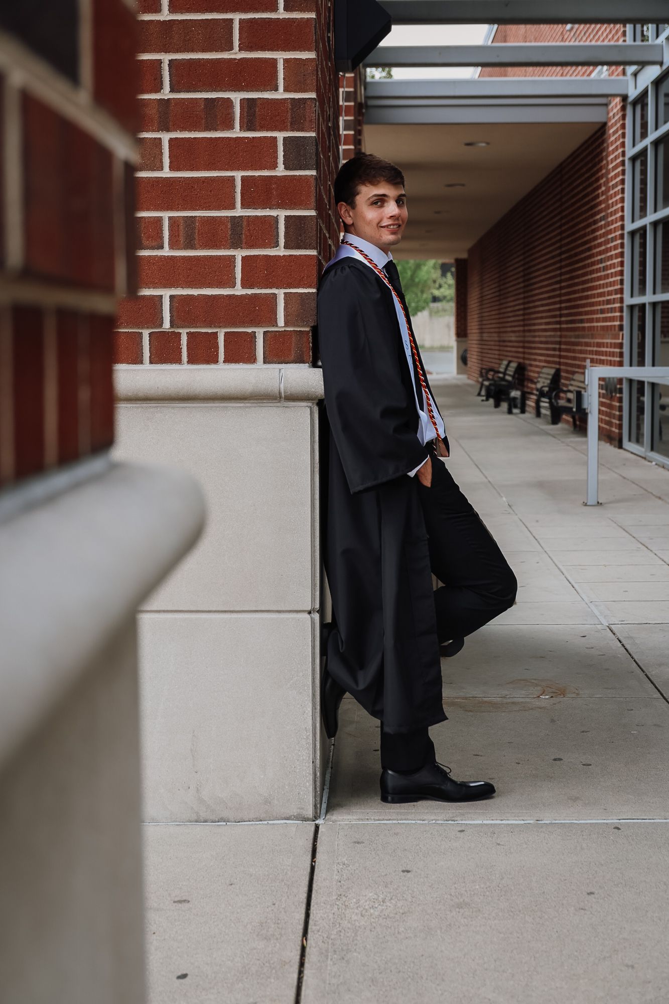 A young man in a black graduation gown leans against a brick building wall, smiling confidently on a school sidewalk.