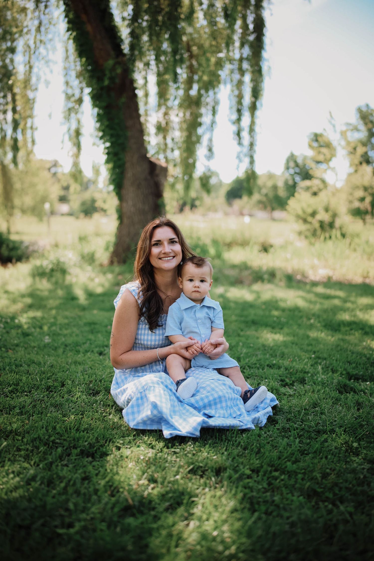 A smiling mother in a blue gingham dress sits on green grass holding her baby boy in a sunny park.