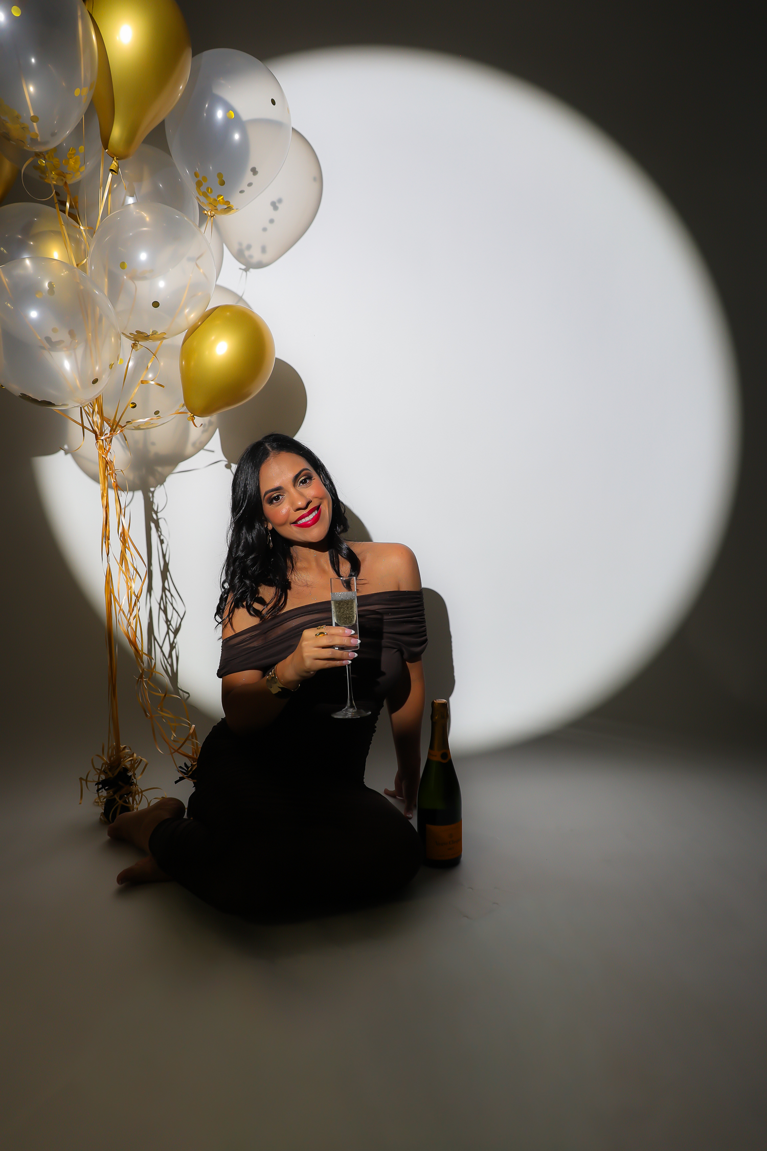 Woman in black dress sitting on floor holding champagne glass with gold and white balloons at a celebration party.