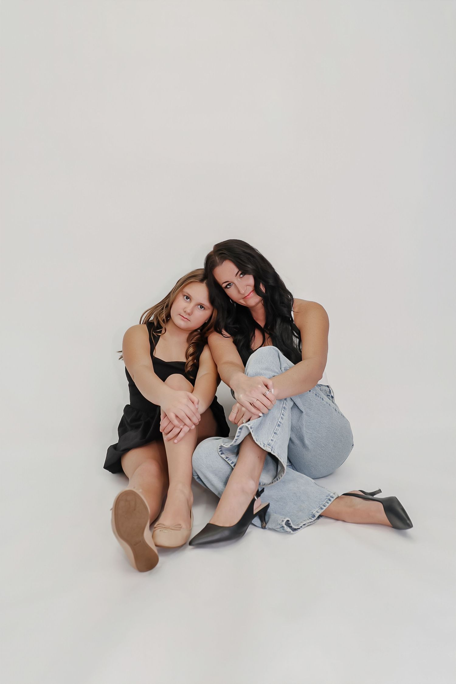Two women sitting together on white background, one in black dress, other in jeans, posing for studio portrait.
