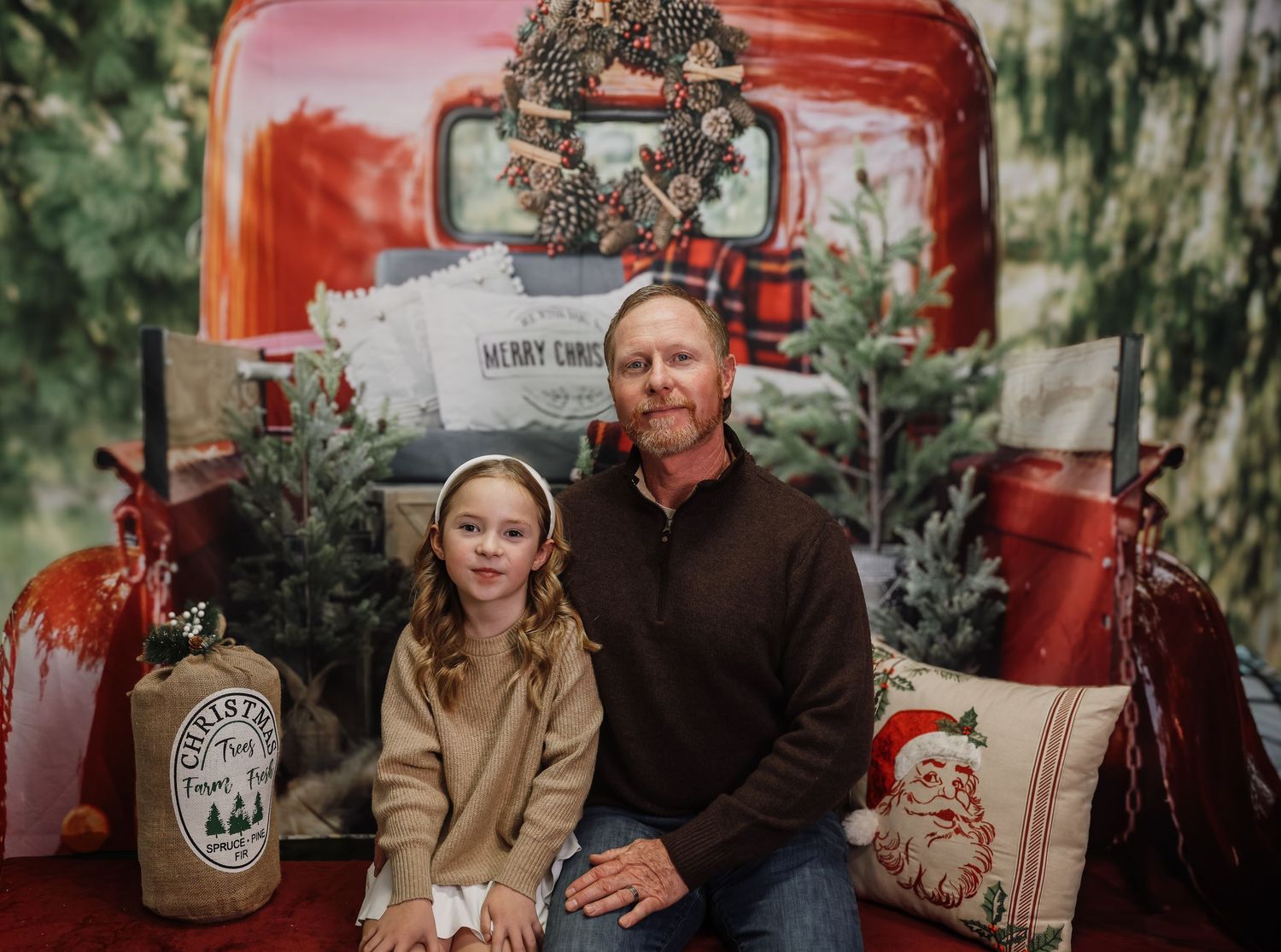 A man and young girl pose for a Christmas portrait in front of a festive red truck backdrop with holiday decorations.