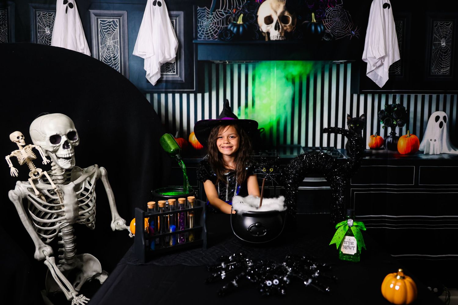 Young girl in witch costume poses with skeleton and Halloween decorations including skulls, ghosts, and a cauldron.
