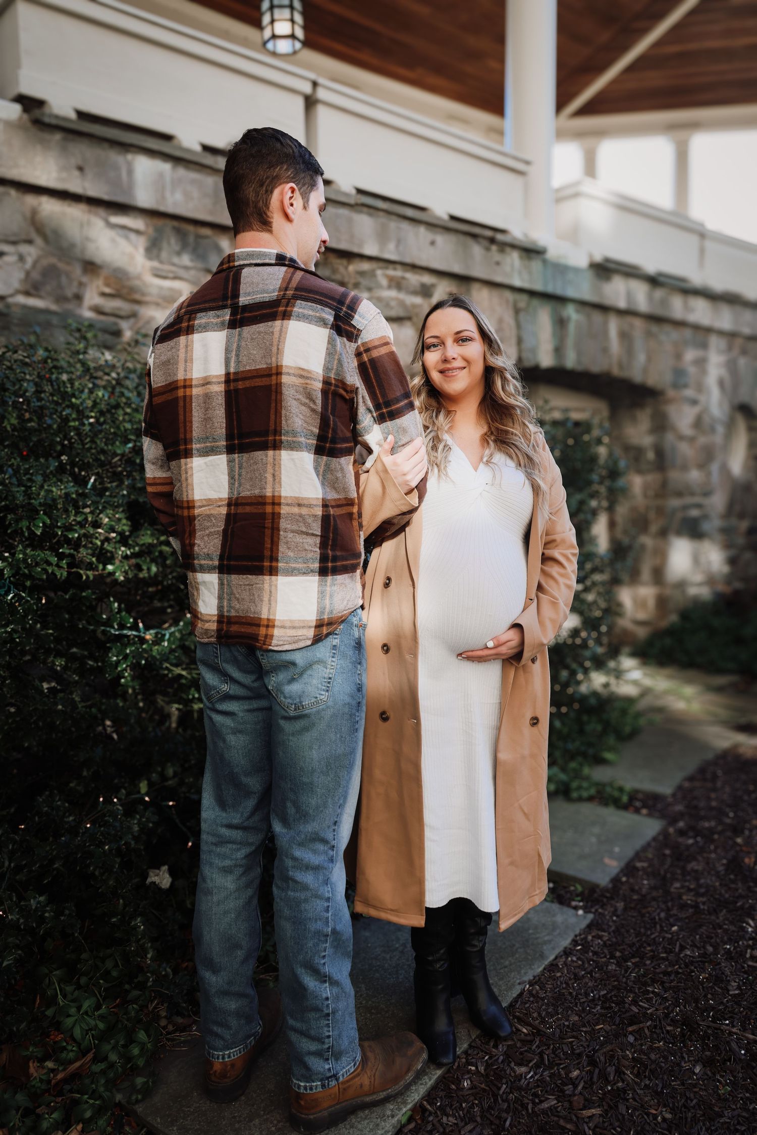 Pregnant woman in white dress and tan coat smiles at partner in plaid shirt outdoors near stone building.