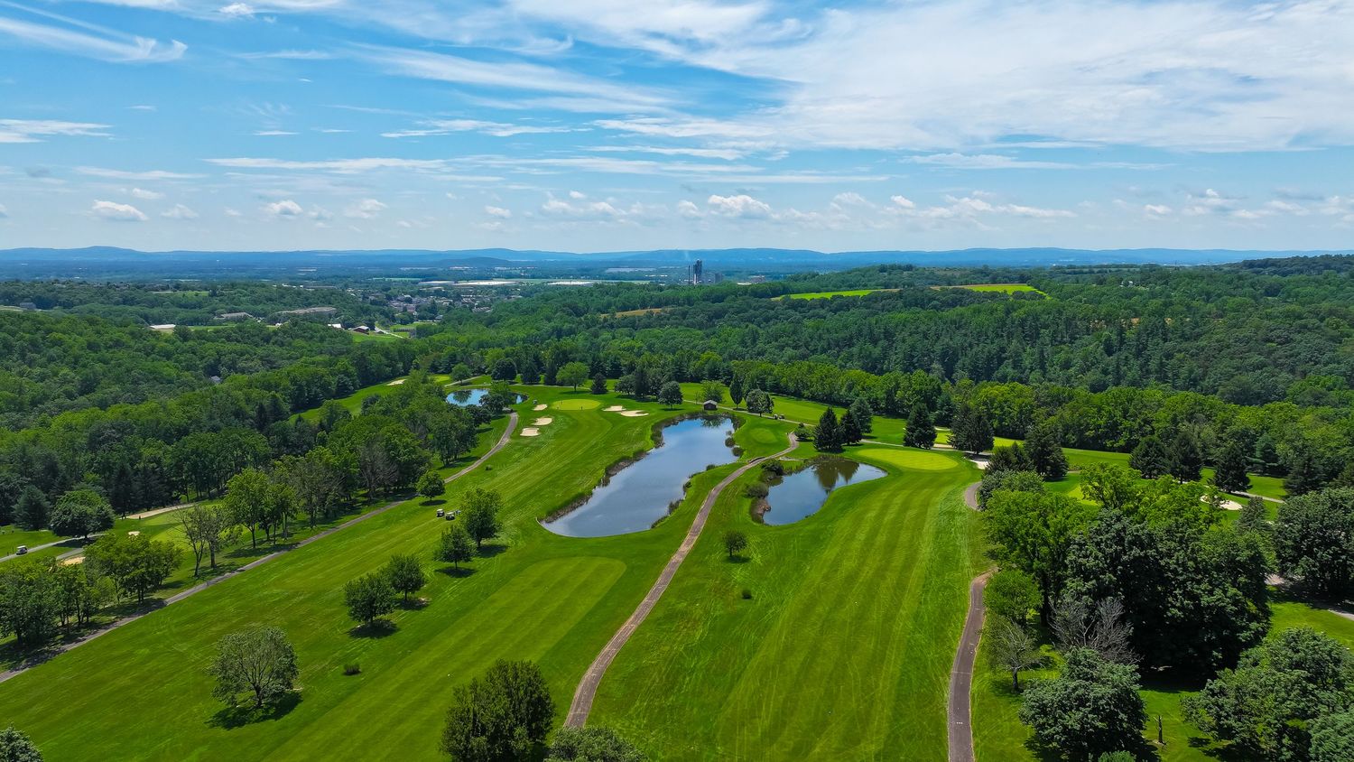 Aerial view of a lush green golf course with ponds, surrounded by dense forests under a bright blue sky.