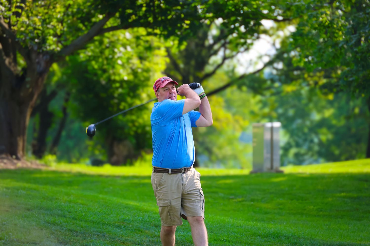 Golfer in khaki shorts and blue shirt takes a swing on a lush green course during a sunny day.