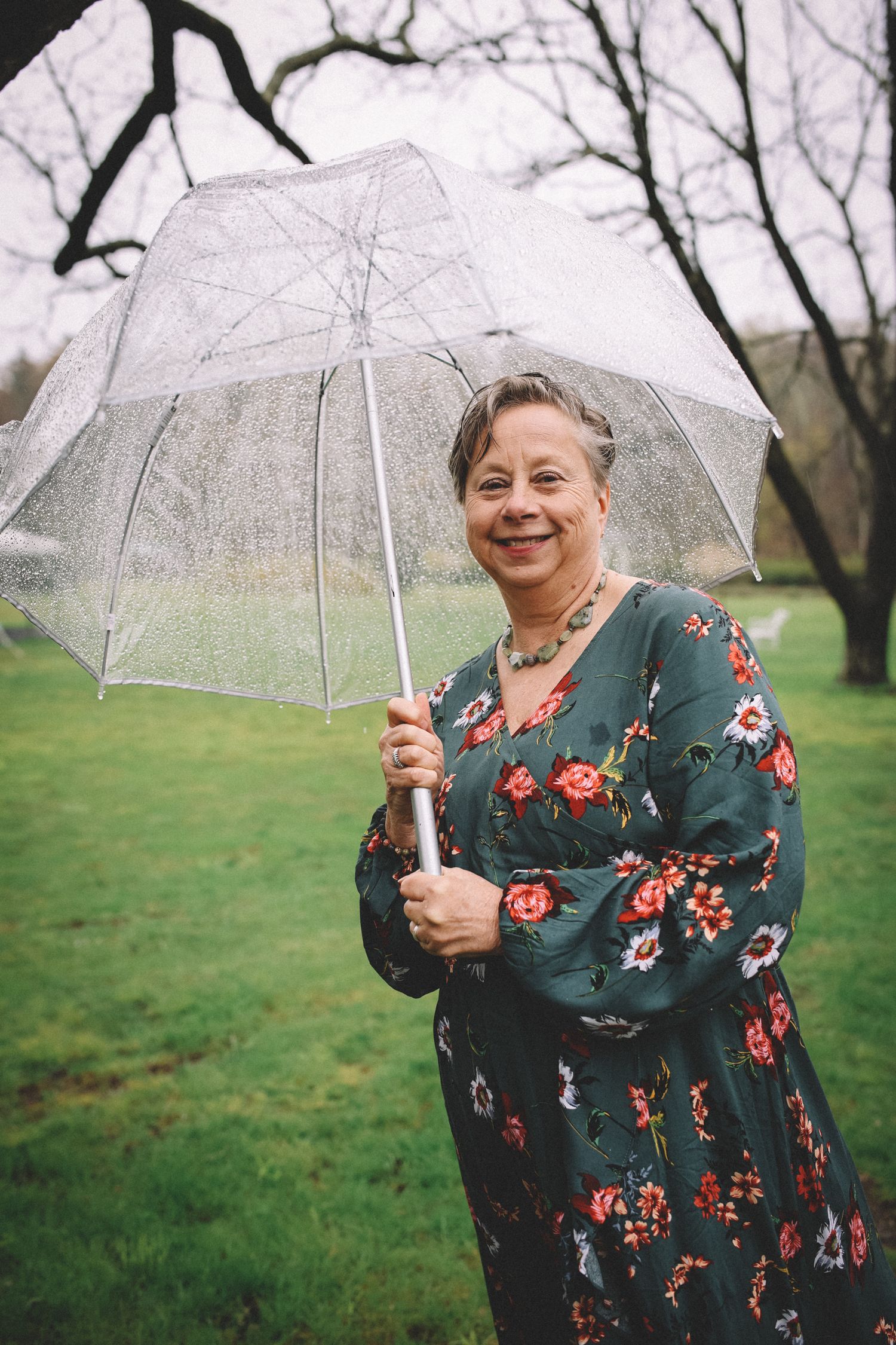A person in a floral dress holds a clear umbrella while standing in a grassy field on an overcast day.