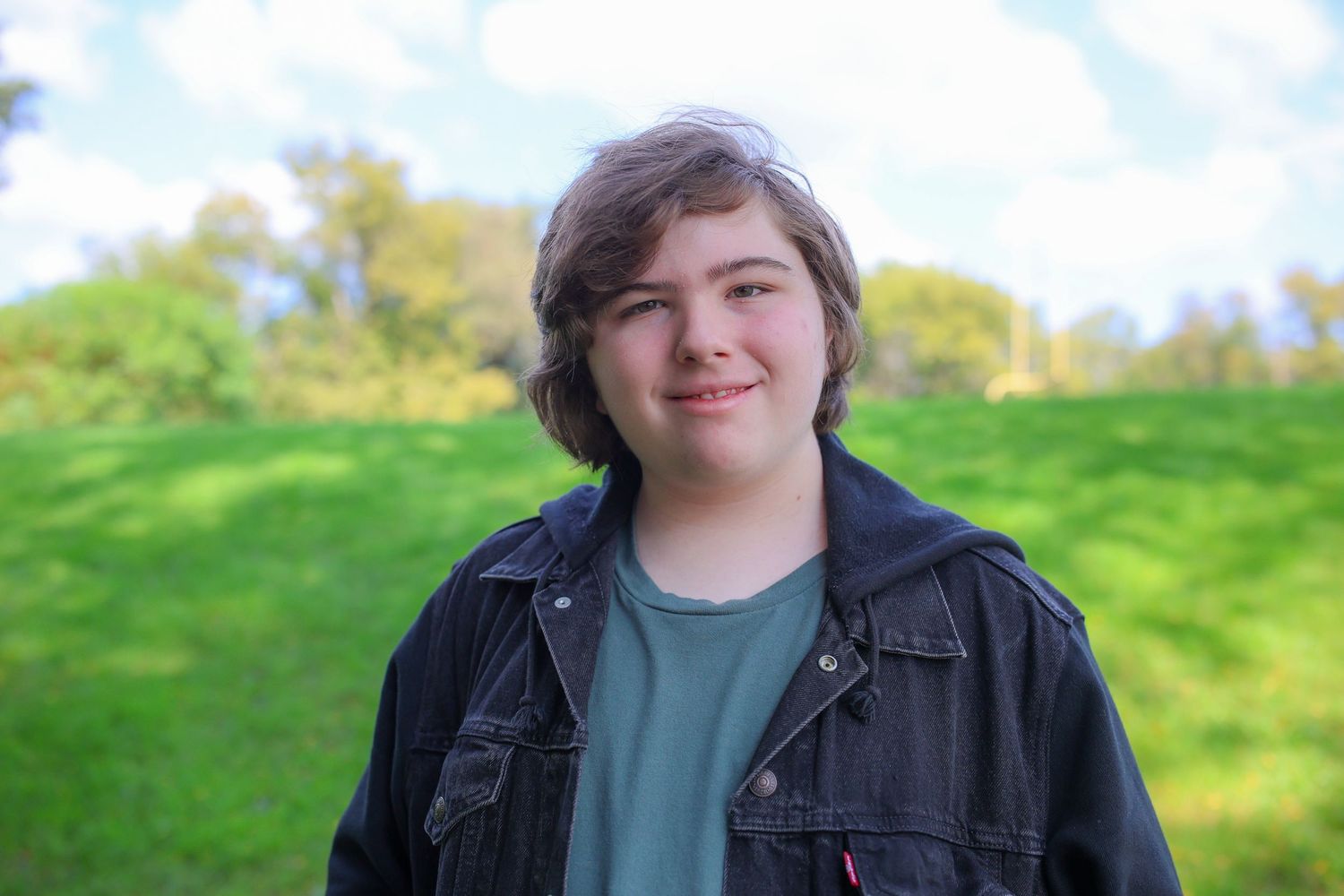 A person wearing a black jacket and gray shirt smiles outdoors against a green natural background.