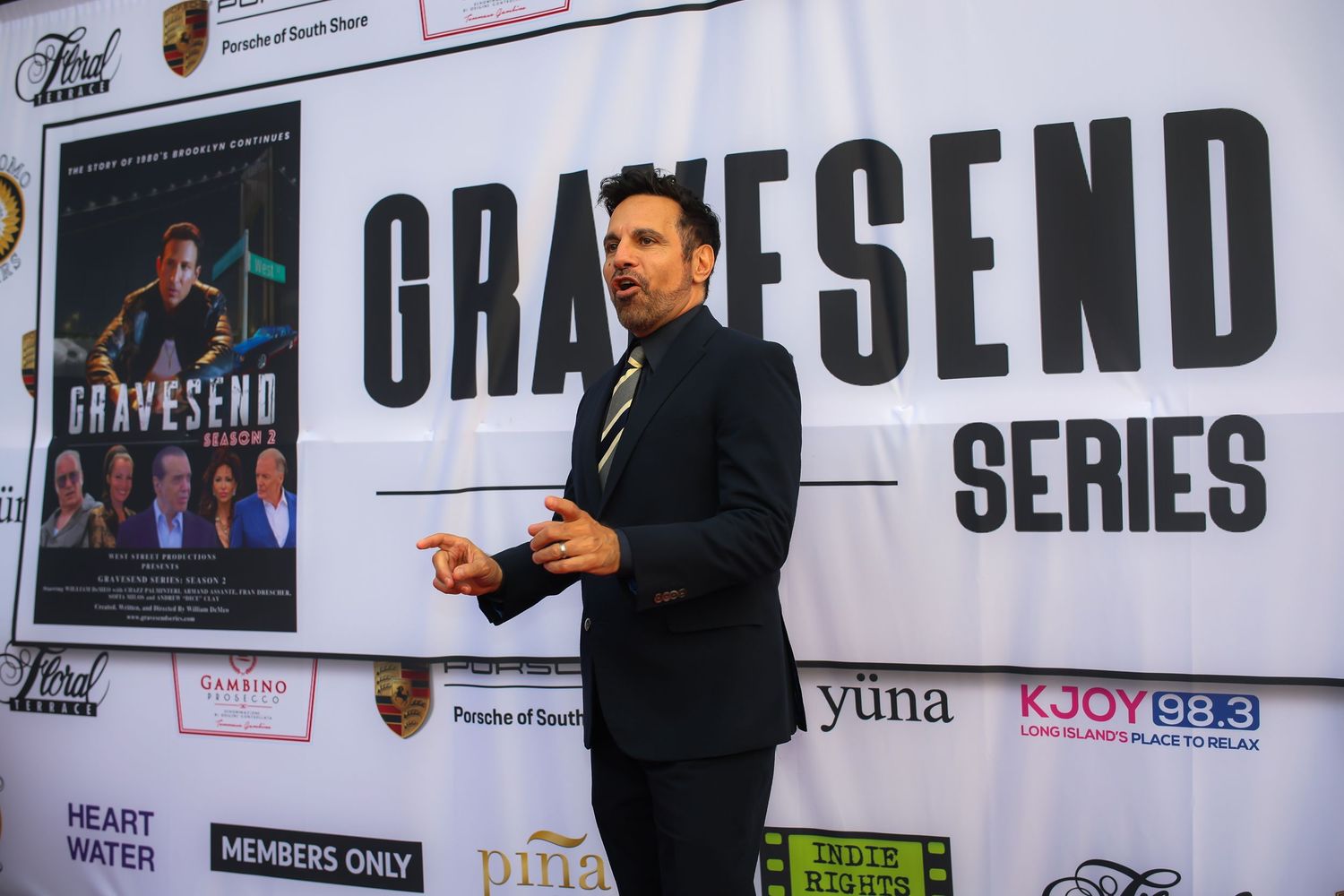 A speaker in a dark suit gestures while presenting on stage with a large 'Gravesend Series' banner behind them.