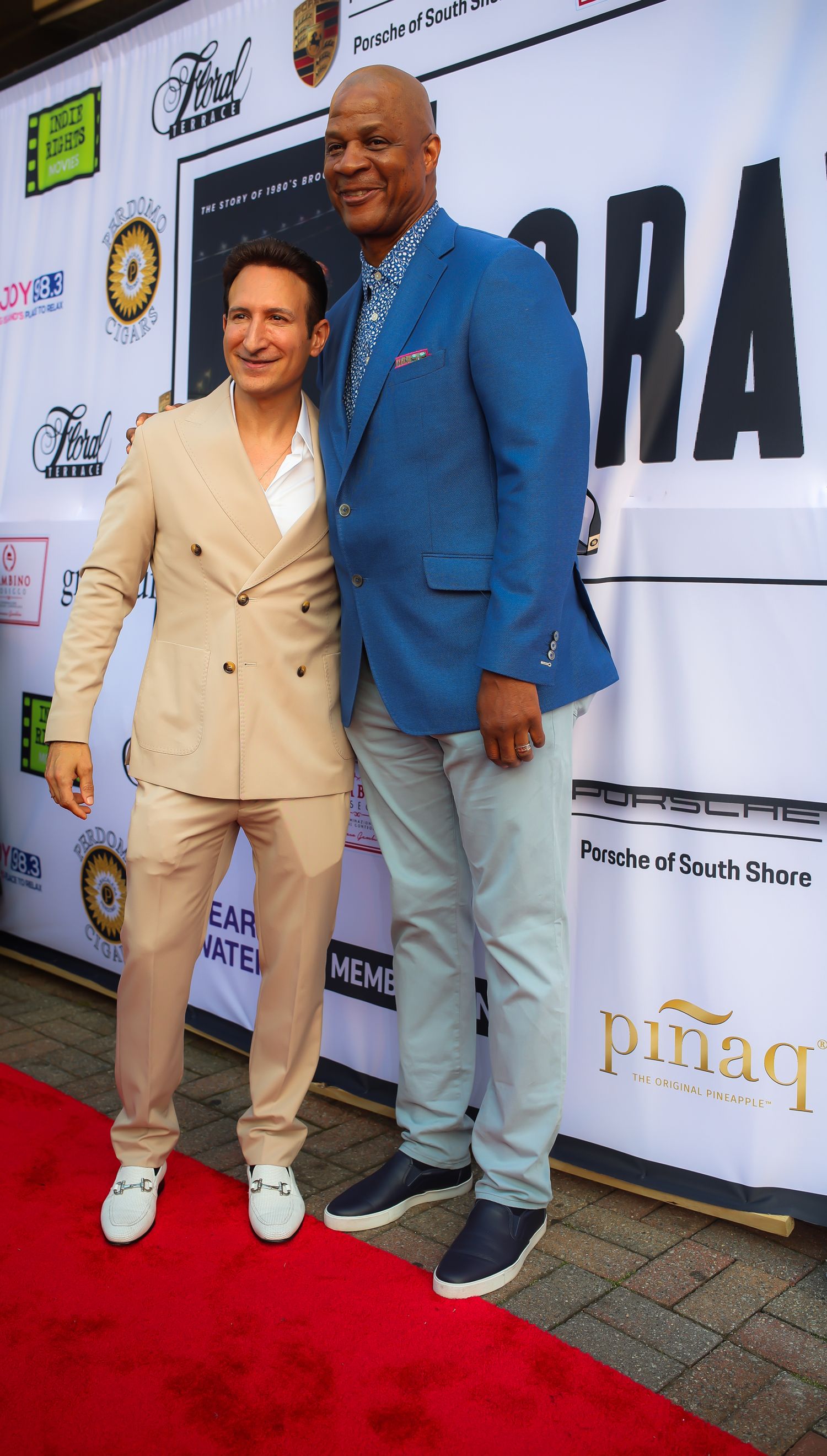 Two people in contrasting outfits pose together on a red carpet at an event with sponsor logos visible in background.