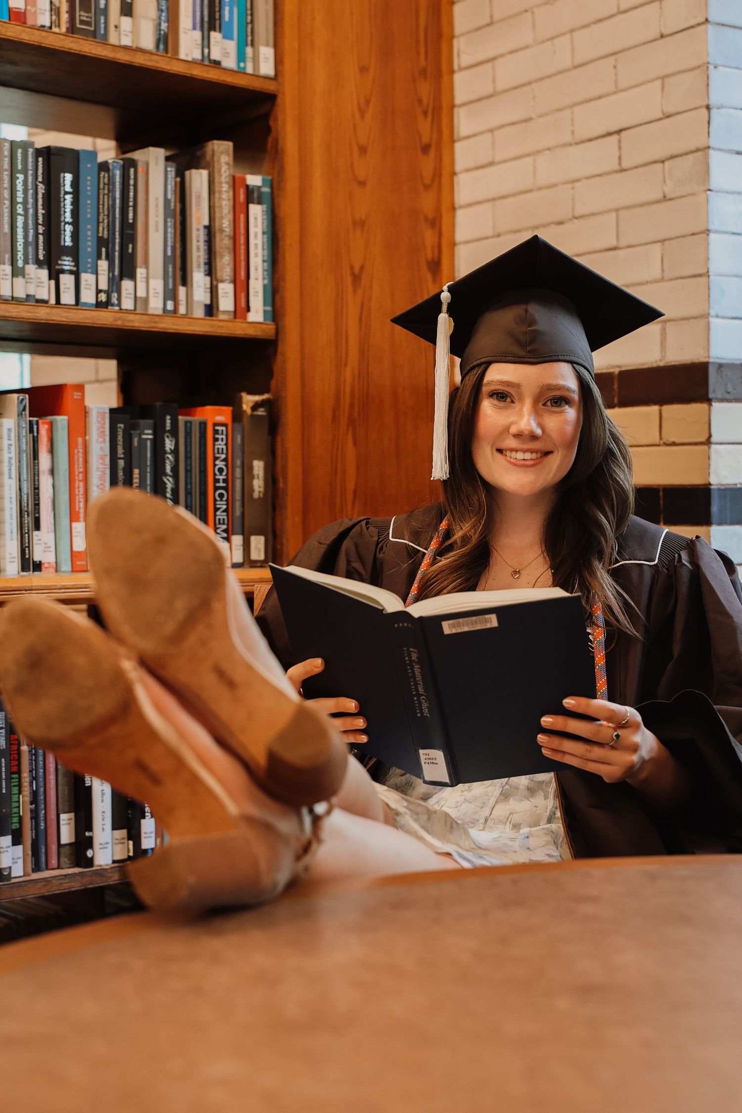 Graduate students in caps and gowns study together at a library table surrounded by wooden bookshelves.