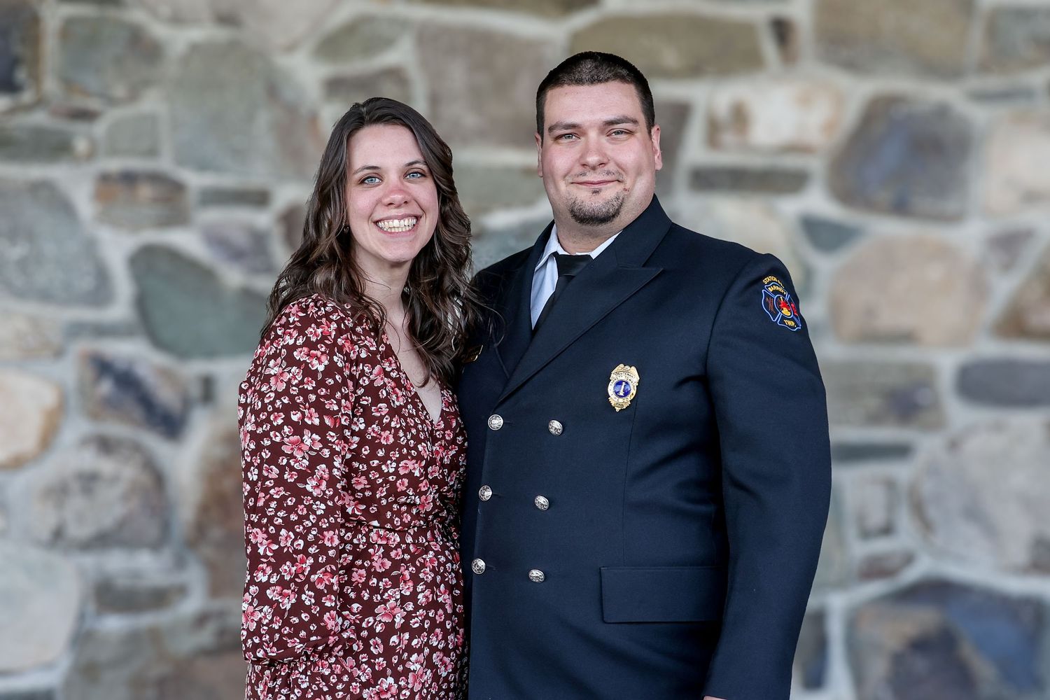 A couple poses together outdoors against a stone wall in coordinated outfits for engagement or portrait photos.