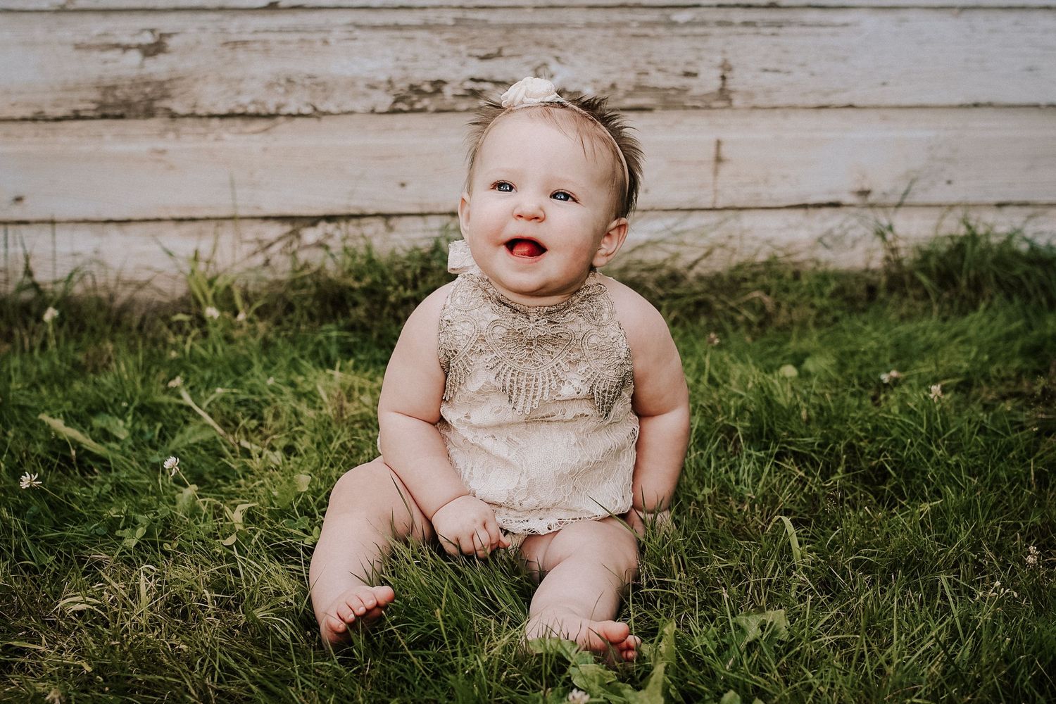 A baby wearing a sparkly gold romper sits on grass in front of a rustic wooden wall.