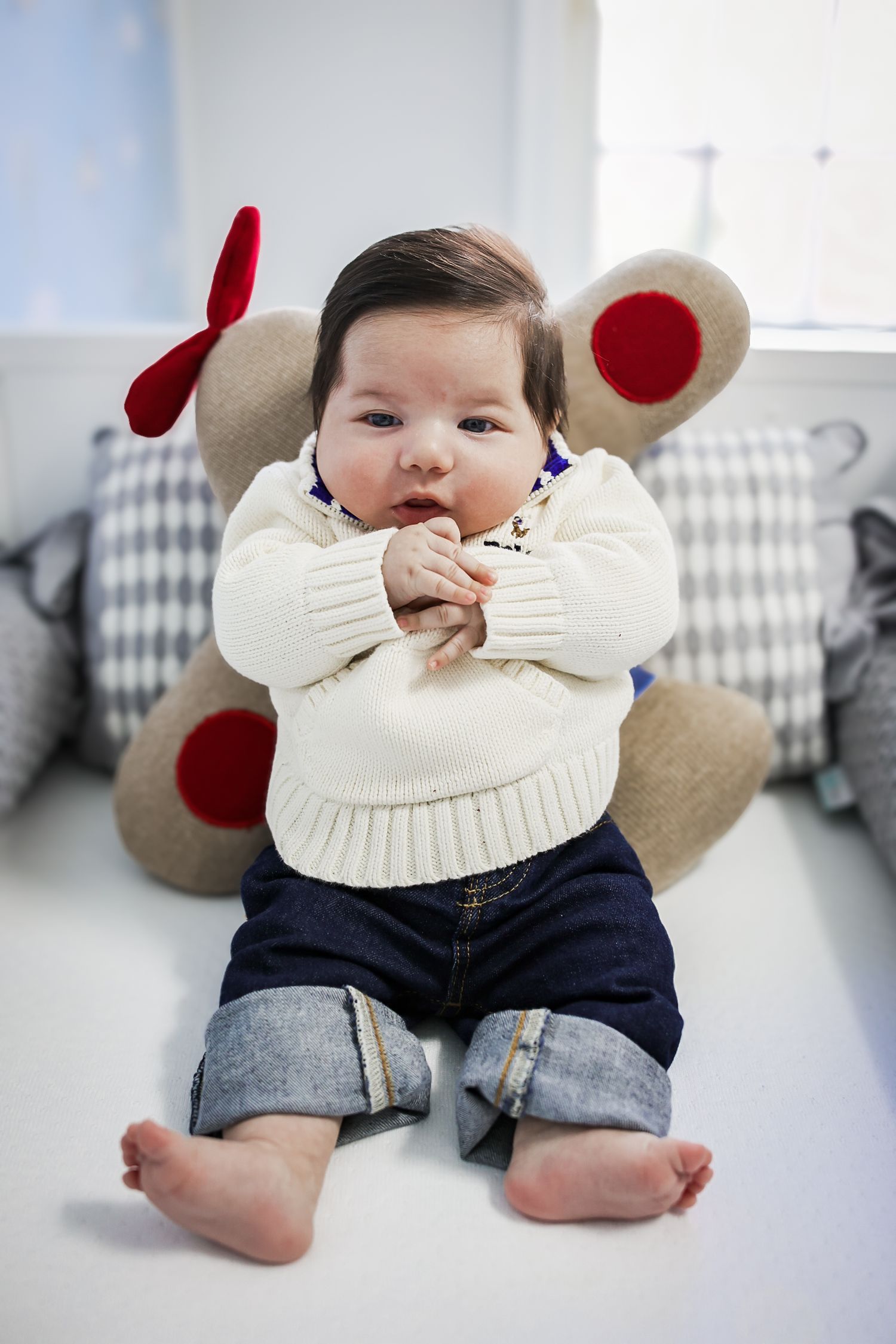 A baby wearing a white sweater and denim shorts lies on a grey and white surface with plush animal ears.