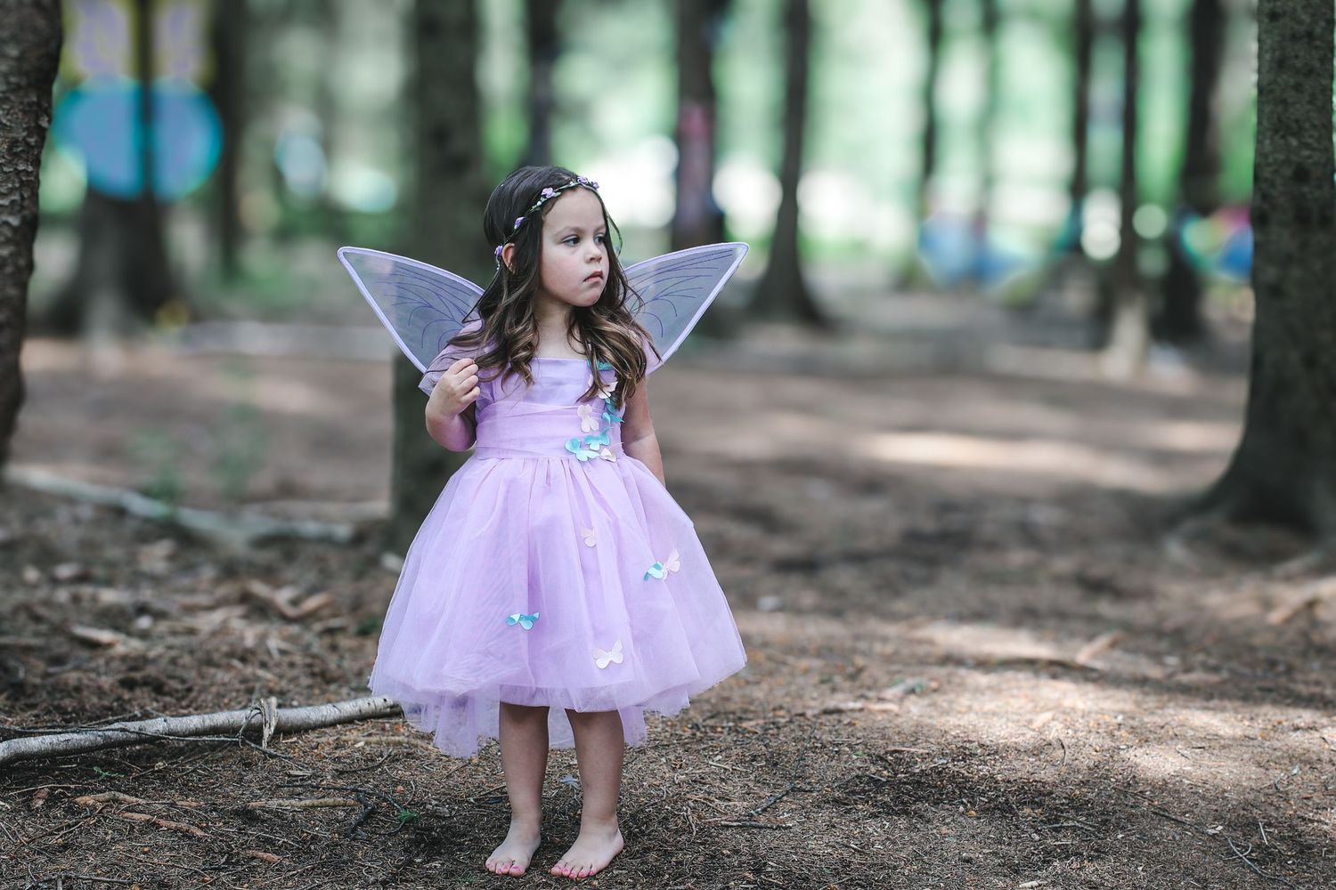 A young child in a pink tulle fairy dress with wings plays in a sunlit forest setting.