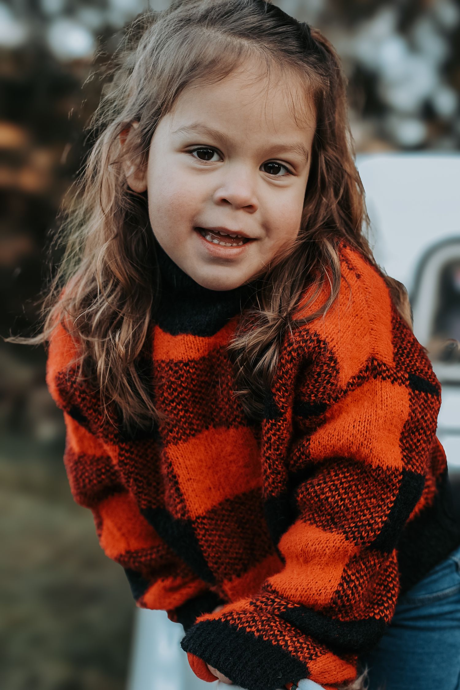 A child wearing a red and black buffalo plaid sweater smiles while playing outdoors.