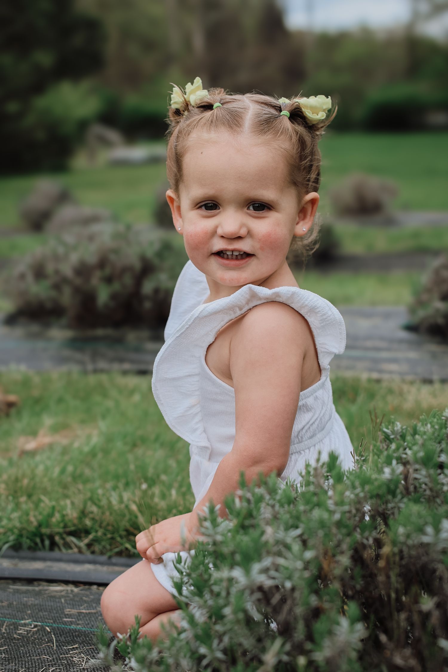 A toddler in a white ruffled dress plays in a garden setting near a stream on a sunny day.