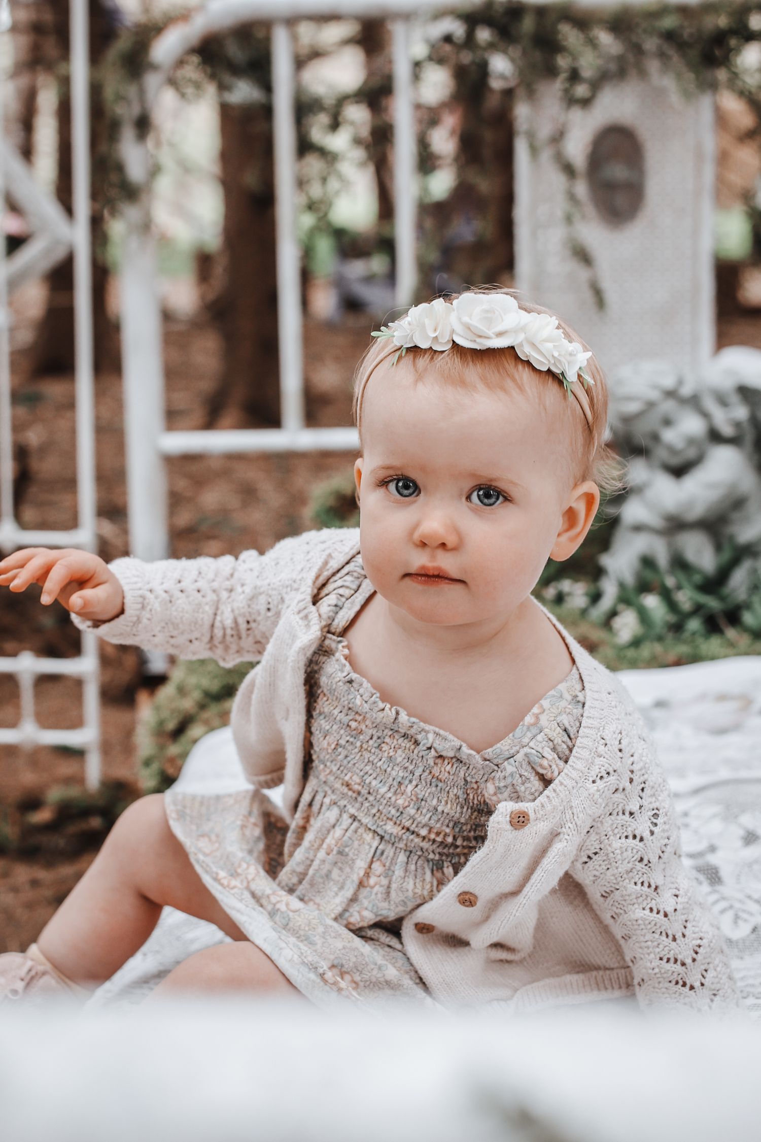 Baby in a white lace dress with floral headband lies on white blanket in outdoor garden setting.
