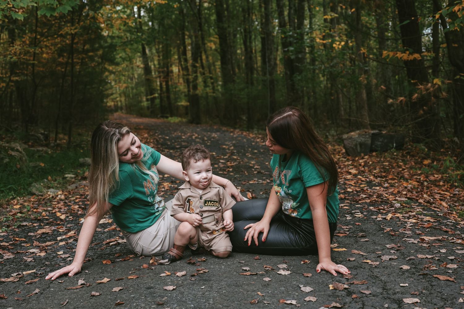 A couple in green shirts playfully poses together on fallen autumn leaves in a forest setting.