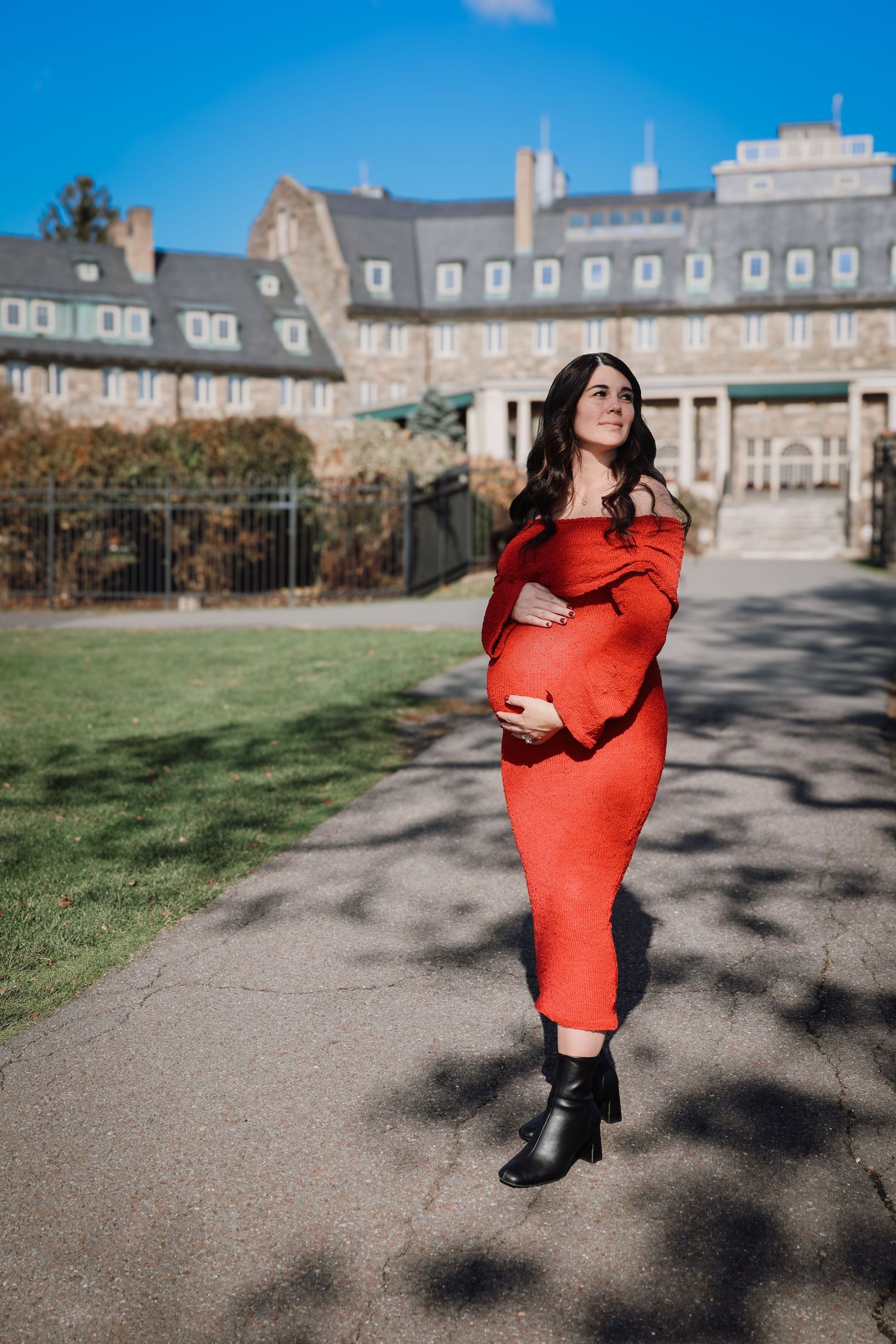 A person in a bright red sweater dress poses on a gravel path with historic stone buildings in the background.