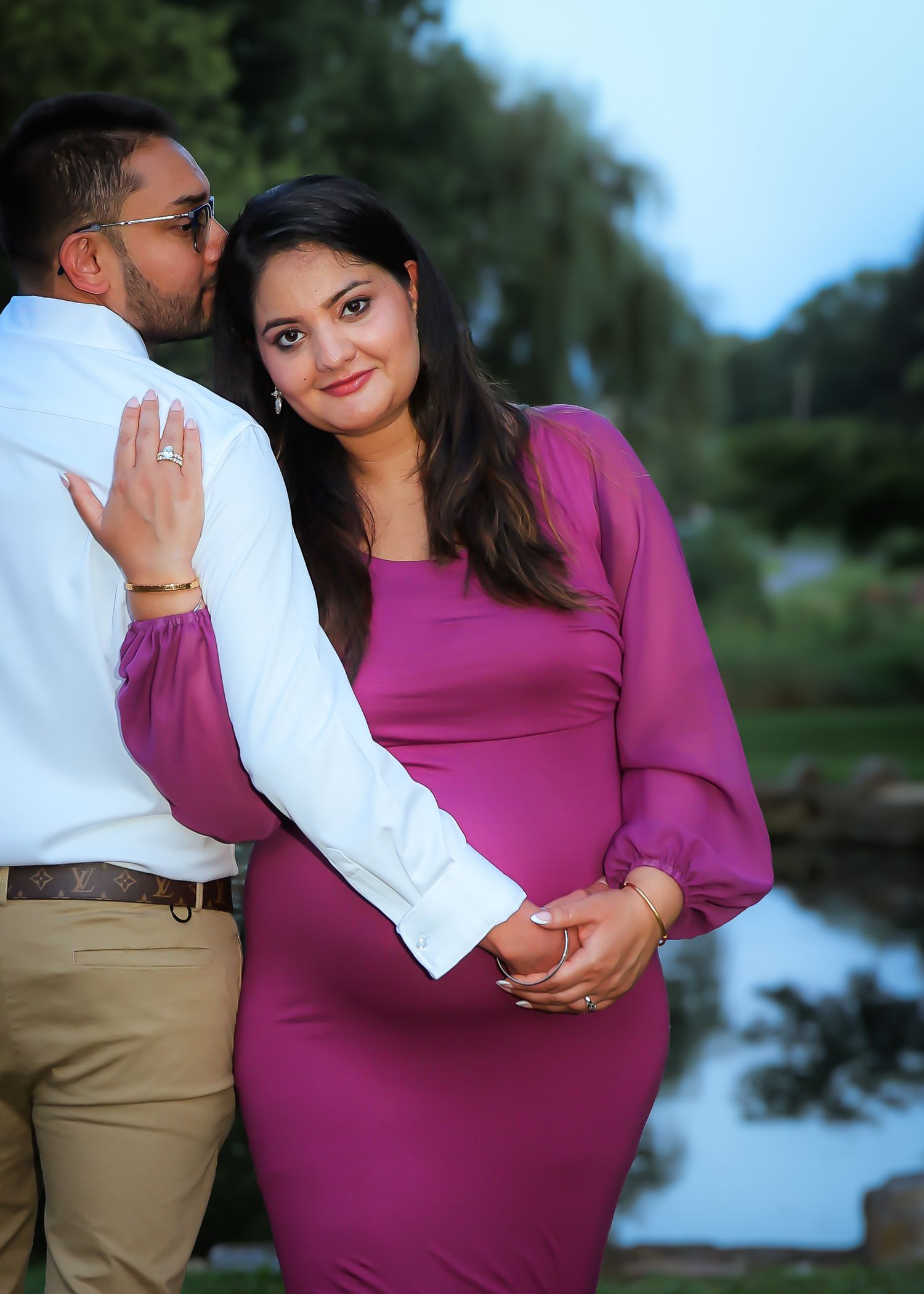 A couple in a magenta dress and white shirt pose together by a lake during an outdoor engagement photoshoot.