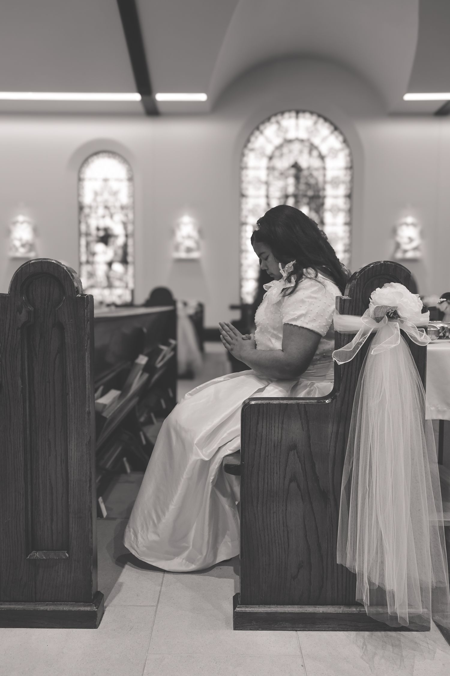 A person in a white wedding dress sits in a church pew decorated with tulle, looking at their phone during a ceremony.