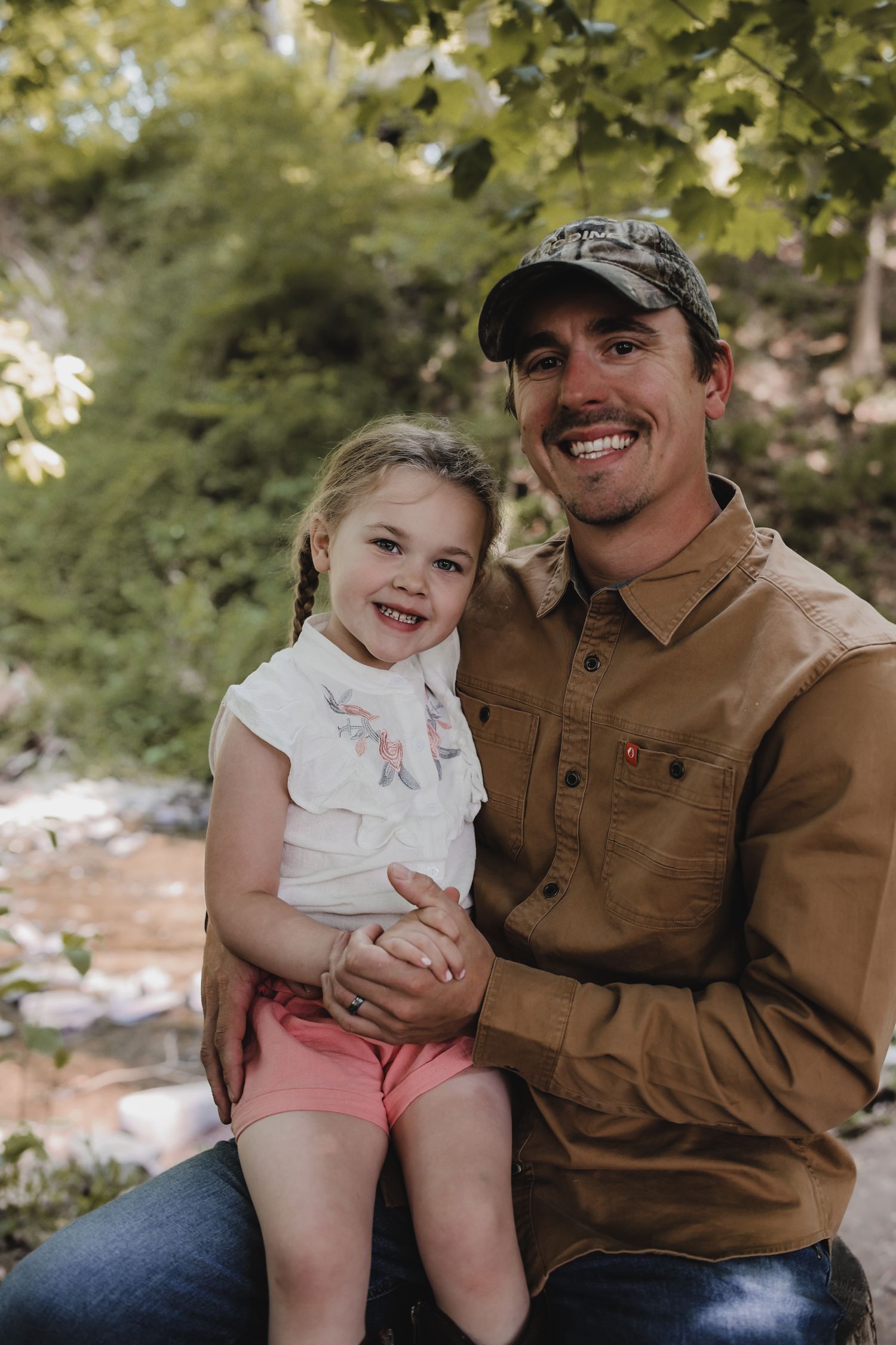 A heartwarming outdoor portrait of a parent and child sitting together on a sunny day among trees and foliage.