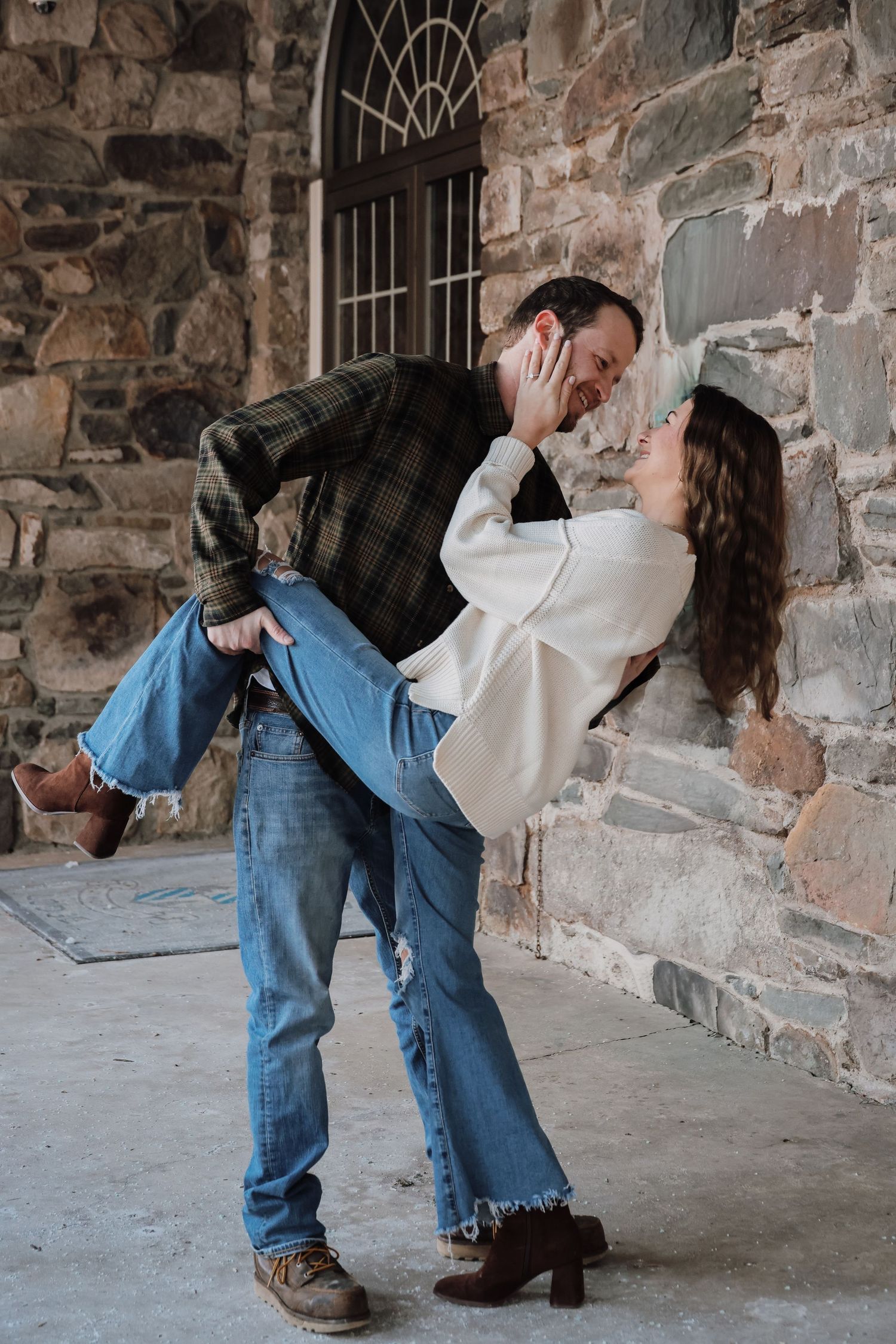 A romantic couple in denim and white share a playful dip pose against a rustic stone wall.