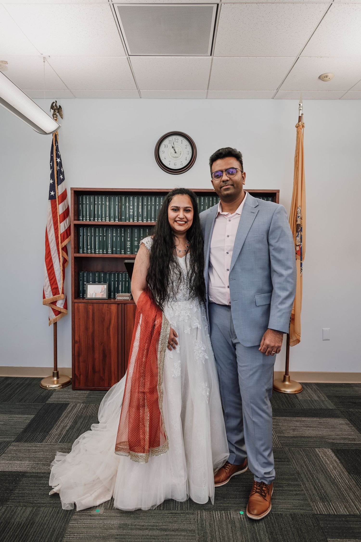 A couple in wedding attire poses in front of a bookshelf and American flag in an office setting.