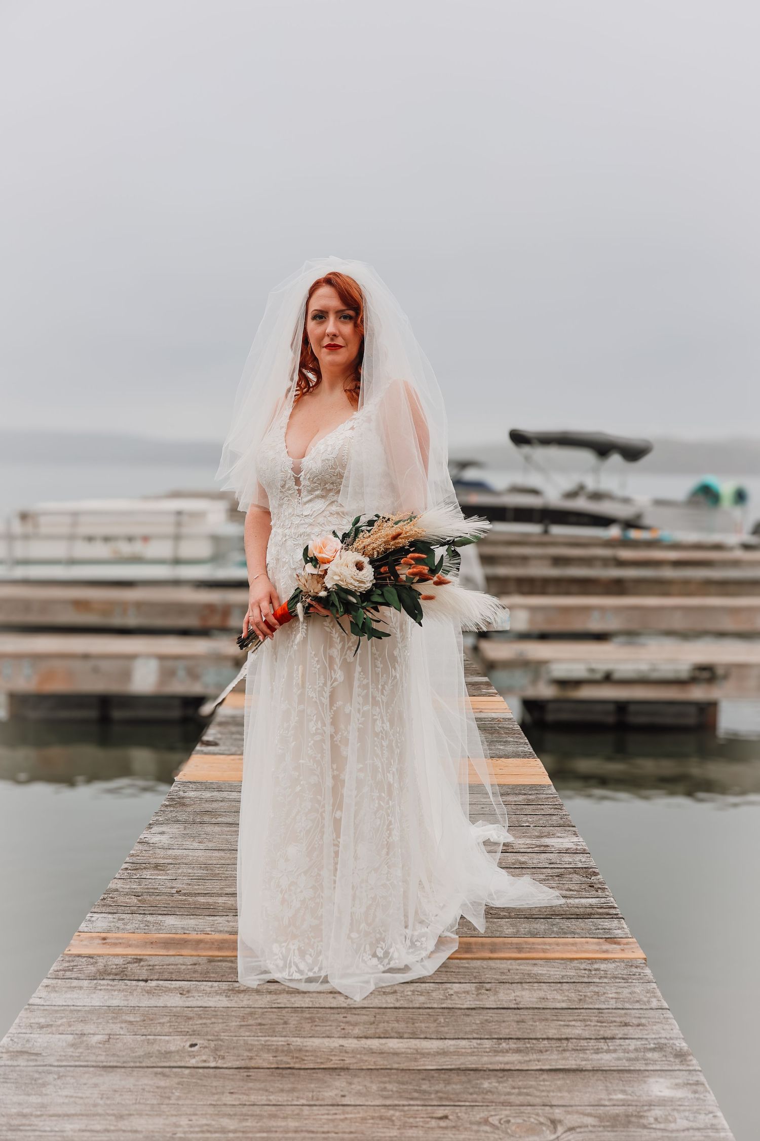 A bride in a lace wedding gown and veil holds a blush and white bouquet while standing on a wooden dock by the water.