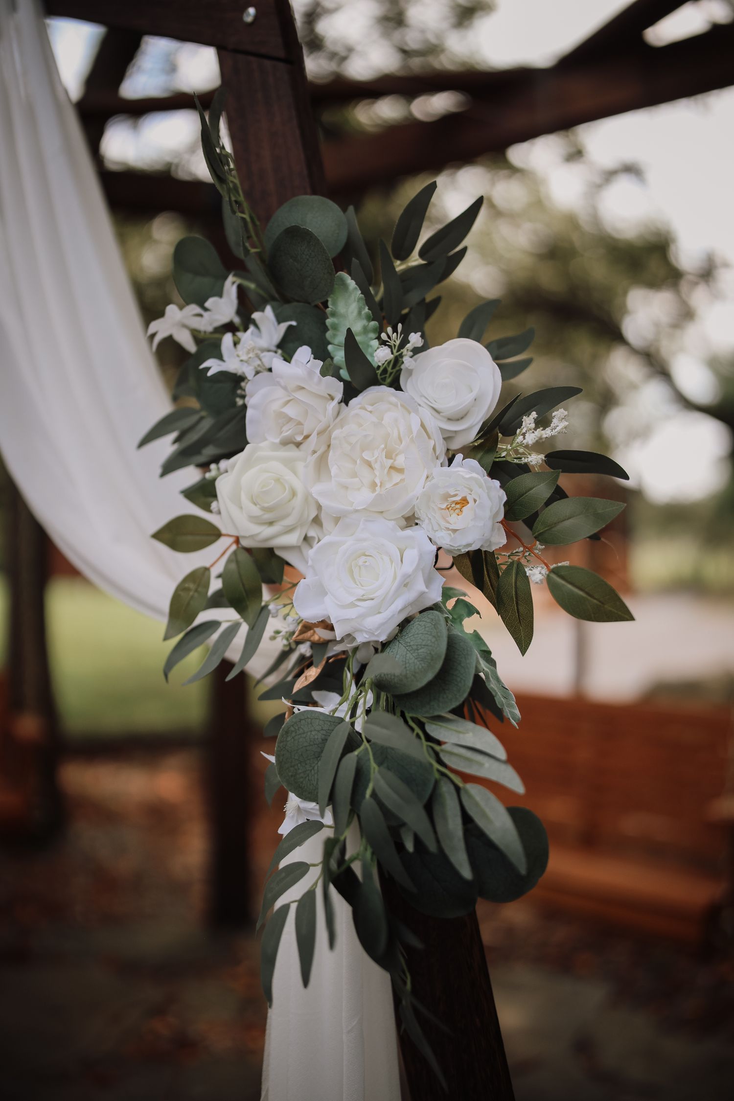 Elegant white rose and eucalyptus floral arrangement adorning a draped wedding arch outdoors.
