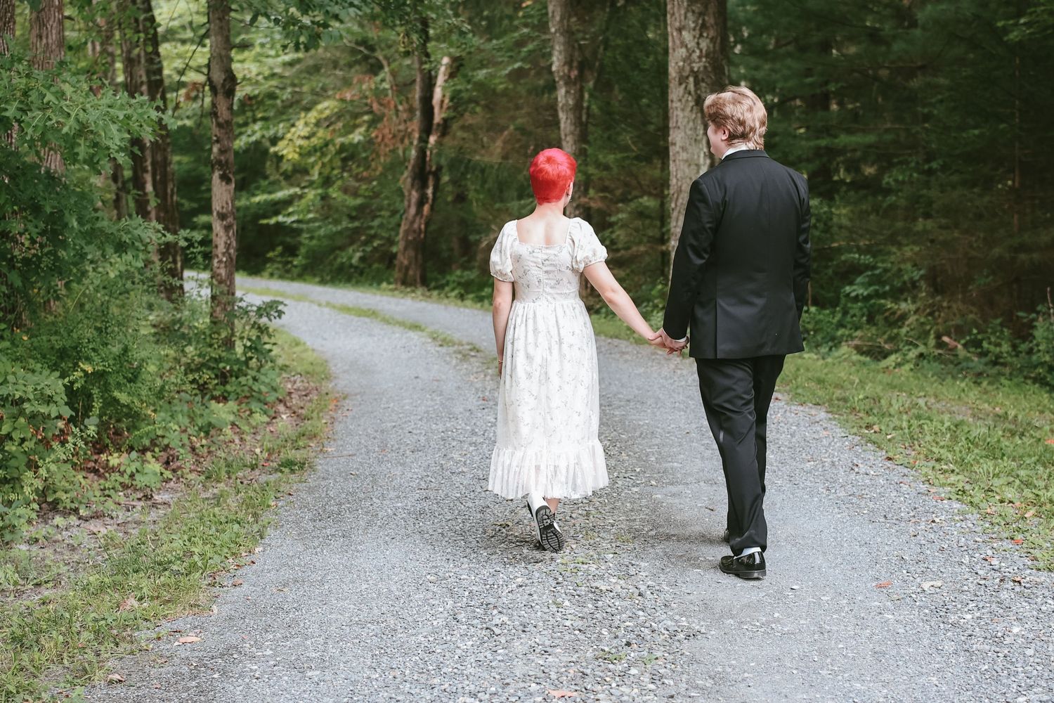 A couple walks away from camera on a gravel path through a lush green forest.