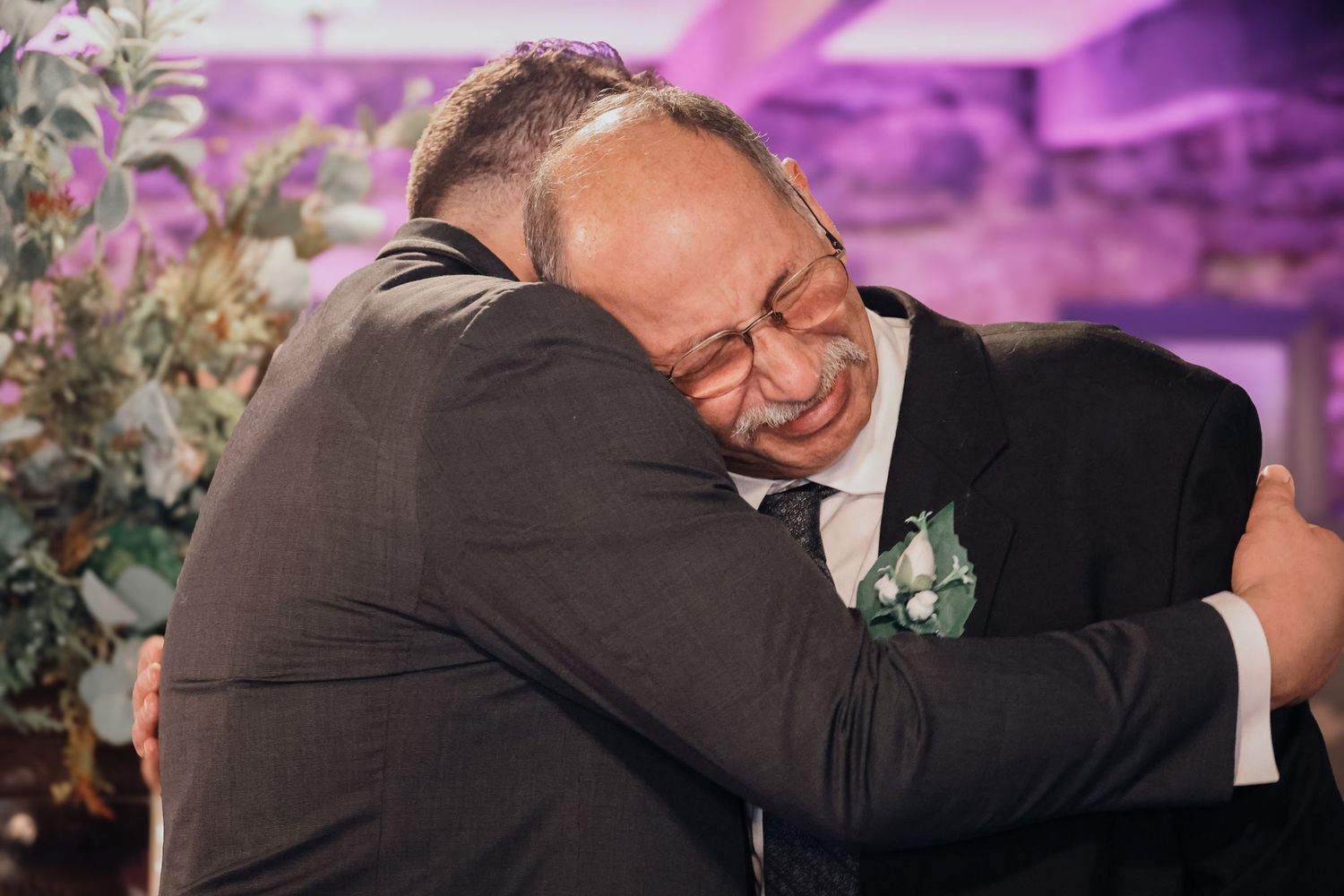 Two people share an emotional embrace at a wedding celebration with purple lighting and floral decorations in the background.
