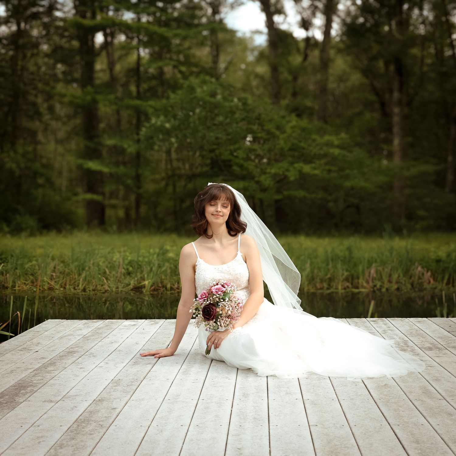 Bride in white wedding dress sitting gracefully on wooden deck surrounded by lush green forest scenery.
