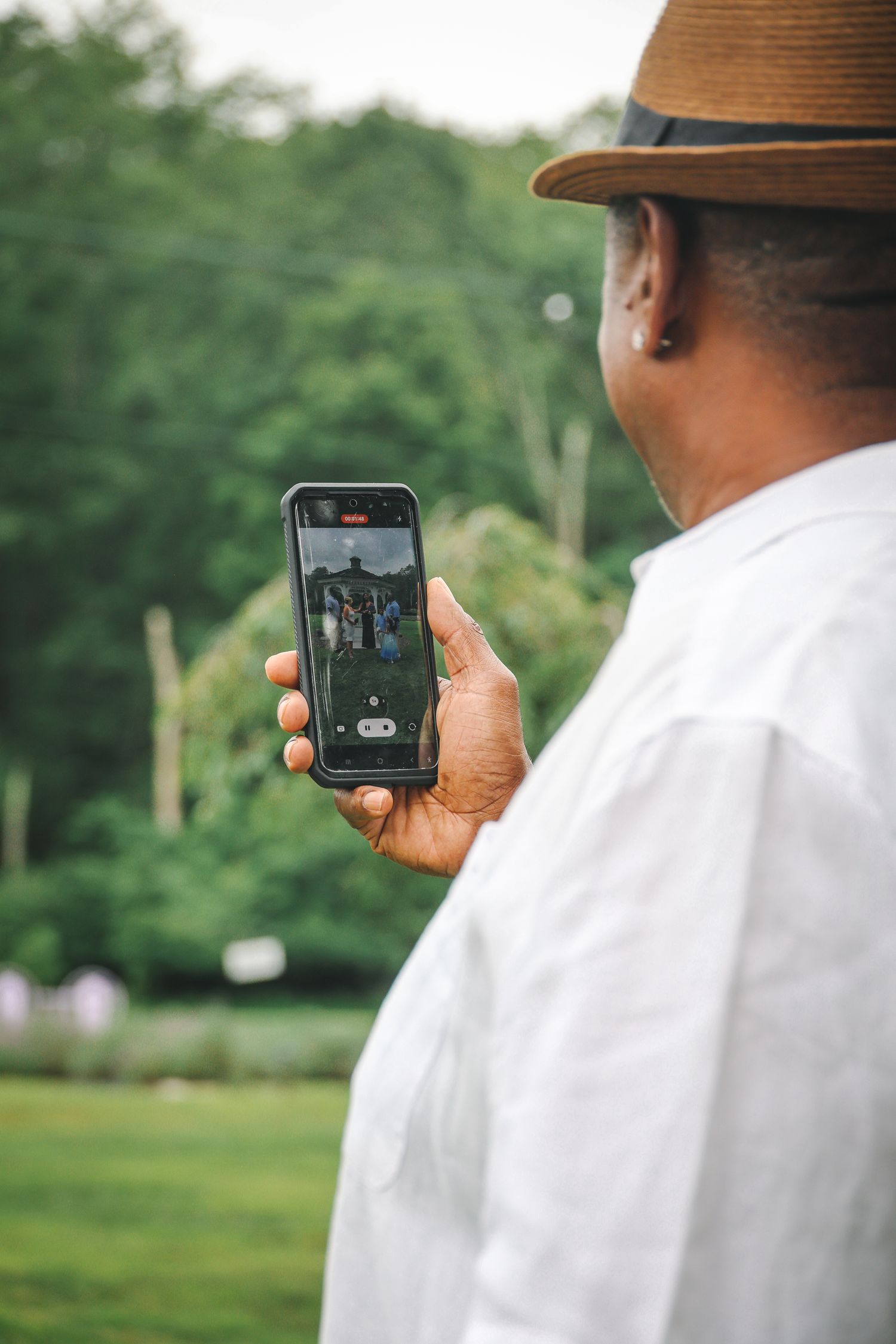 Person in white clothing and brown hat looking at a mobile phone screen outdoors in a green park setting.