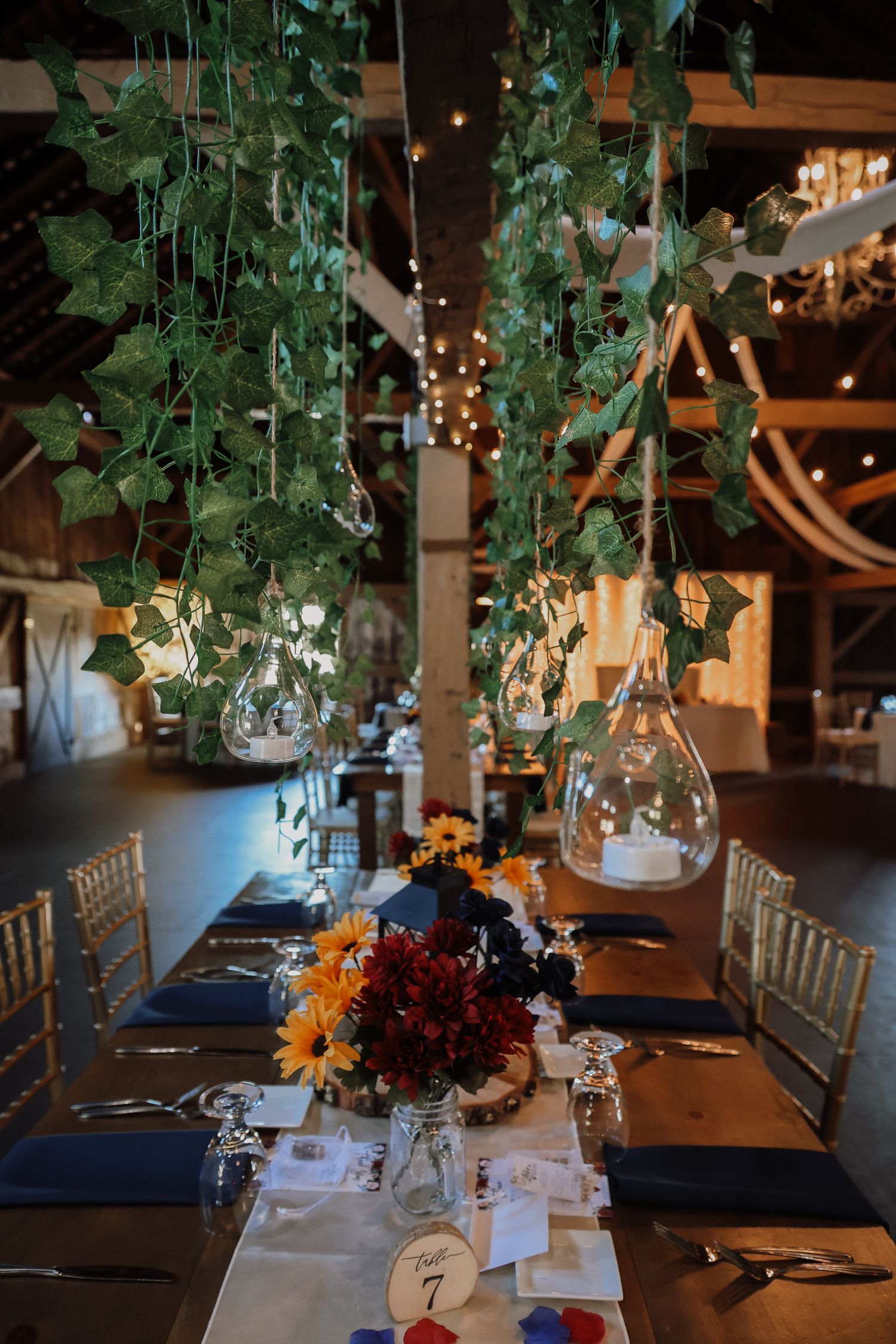 Wedding reception table decorated with hanging greenery, string lights, and autumn floral centerpieces at an indoor venue.