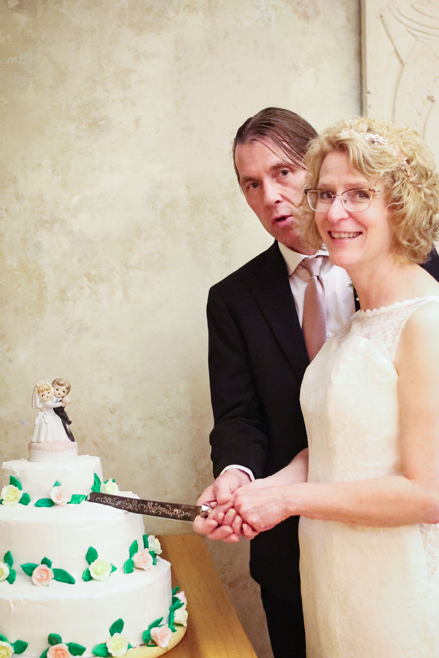 A couple cutting their white tiered wedding cake decorated with green leaves and flowers at their reception.