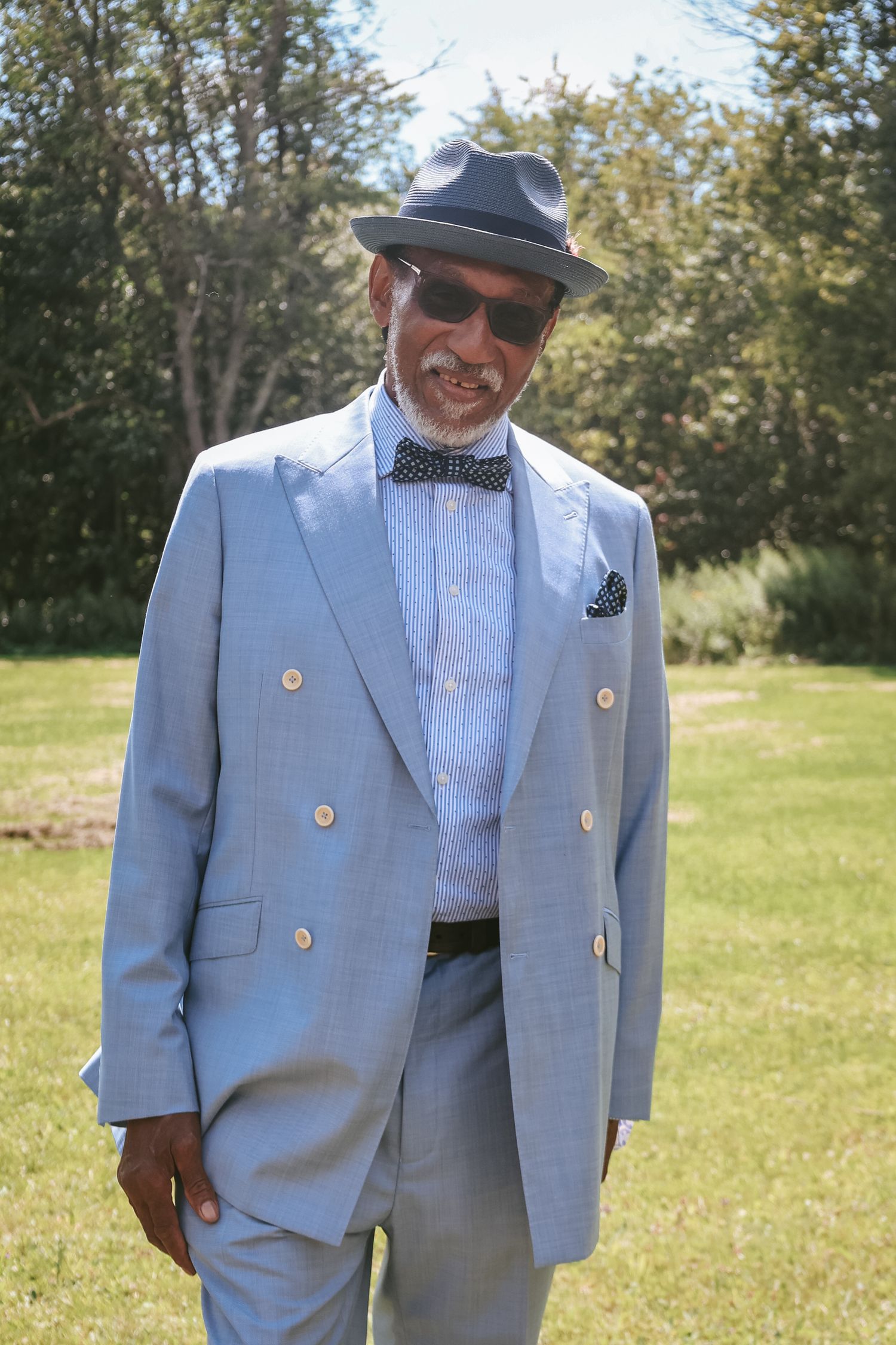 A person wearing a light blue suit with gold buttons and polka dot bowtie poses in a grassy outdoor setting.
