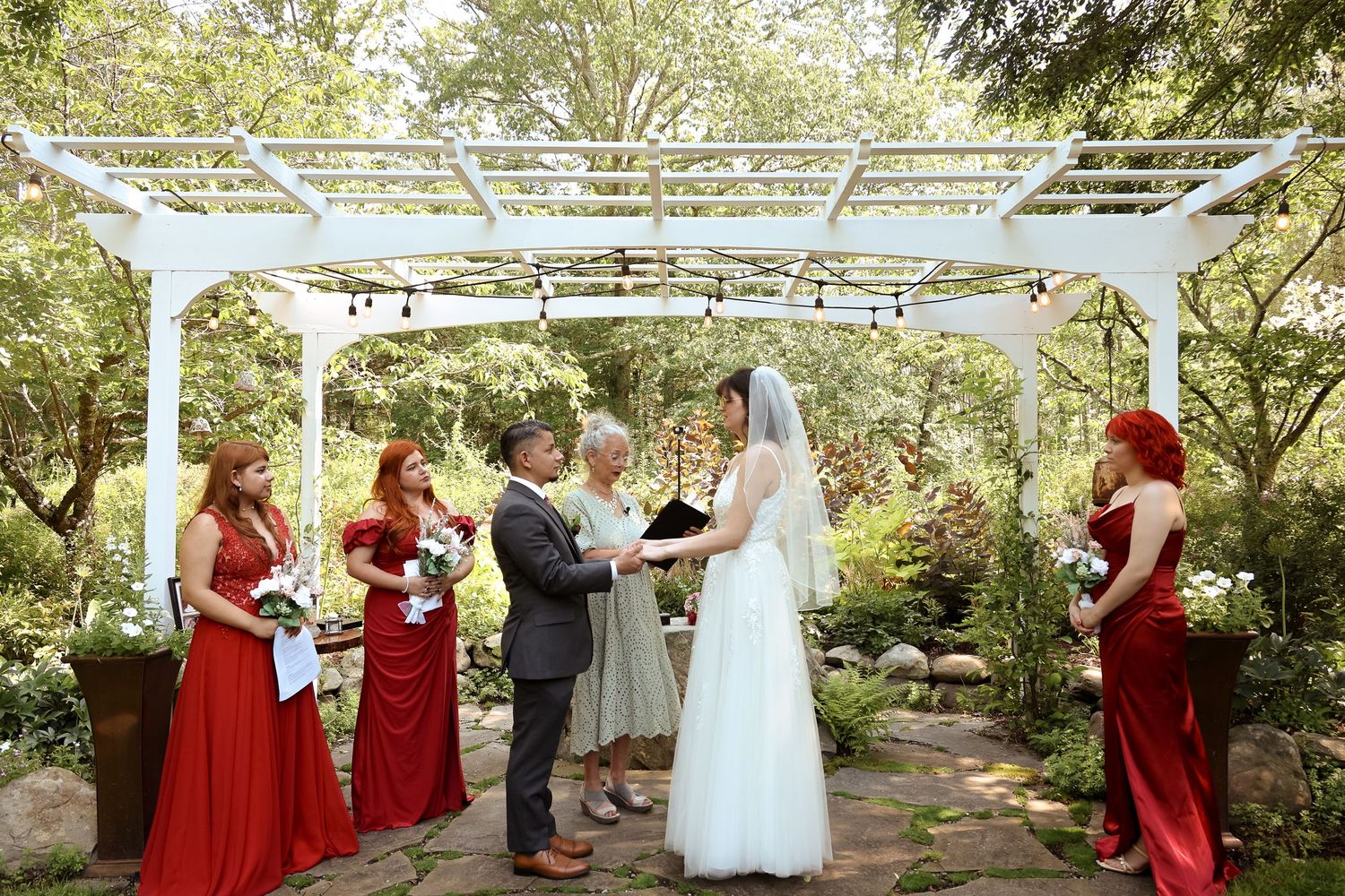An outdoor wedding ceremony takes place under a white pergola with bridesmaids in red dresses standing to the side.