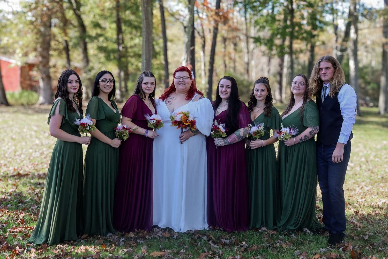 A bridal party poses outdoors wearing alternating green and burgundy floor-length dresses on a sunny autumn day.