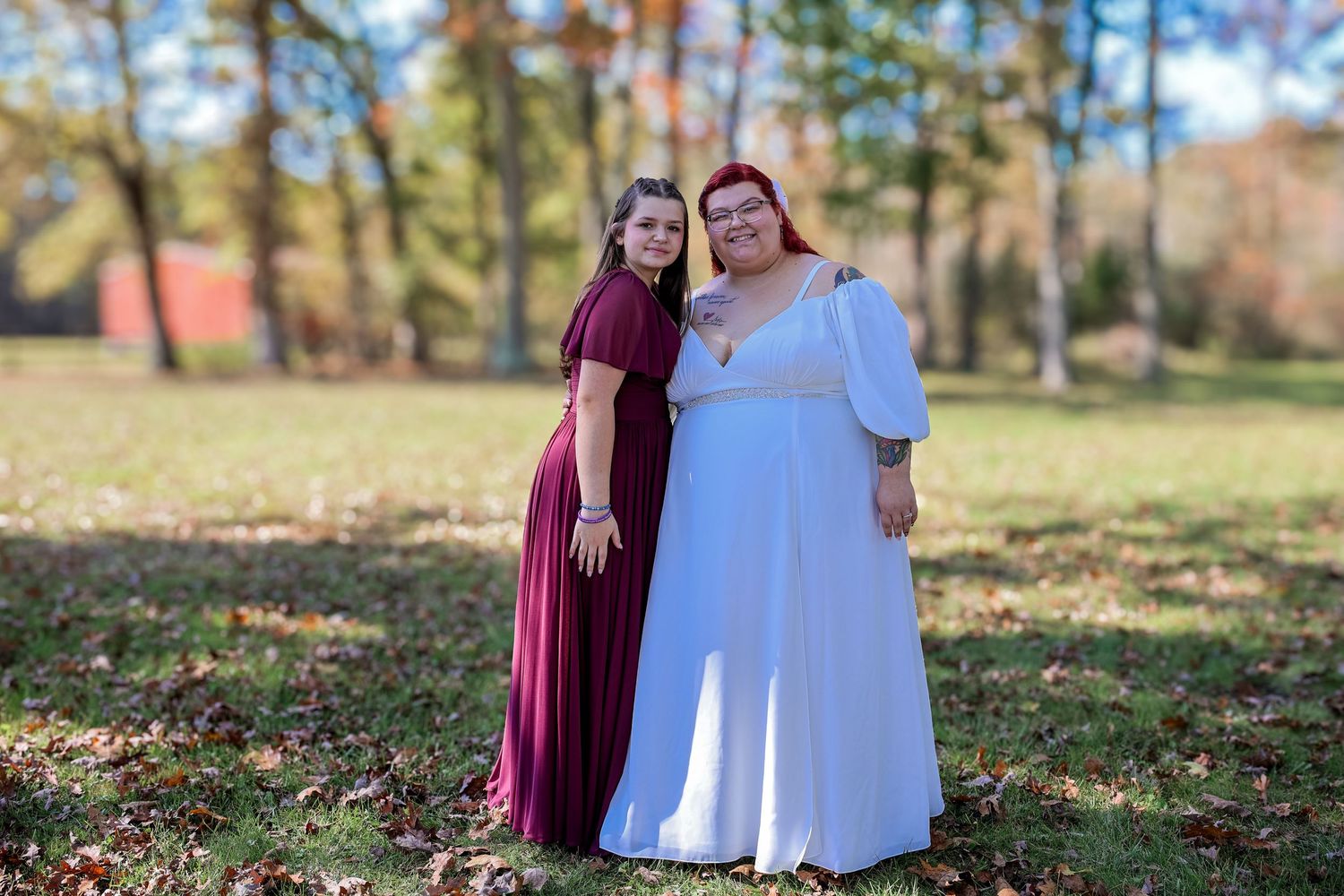 Two people in burgundy and white outfits stand together outdoors on fallen autumn leaves in a park setting.