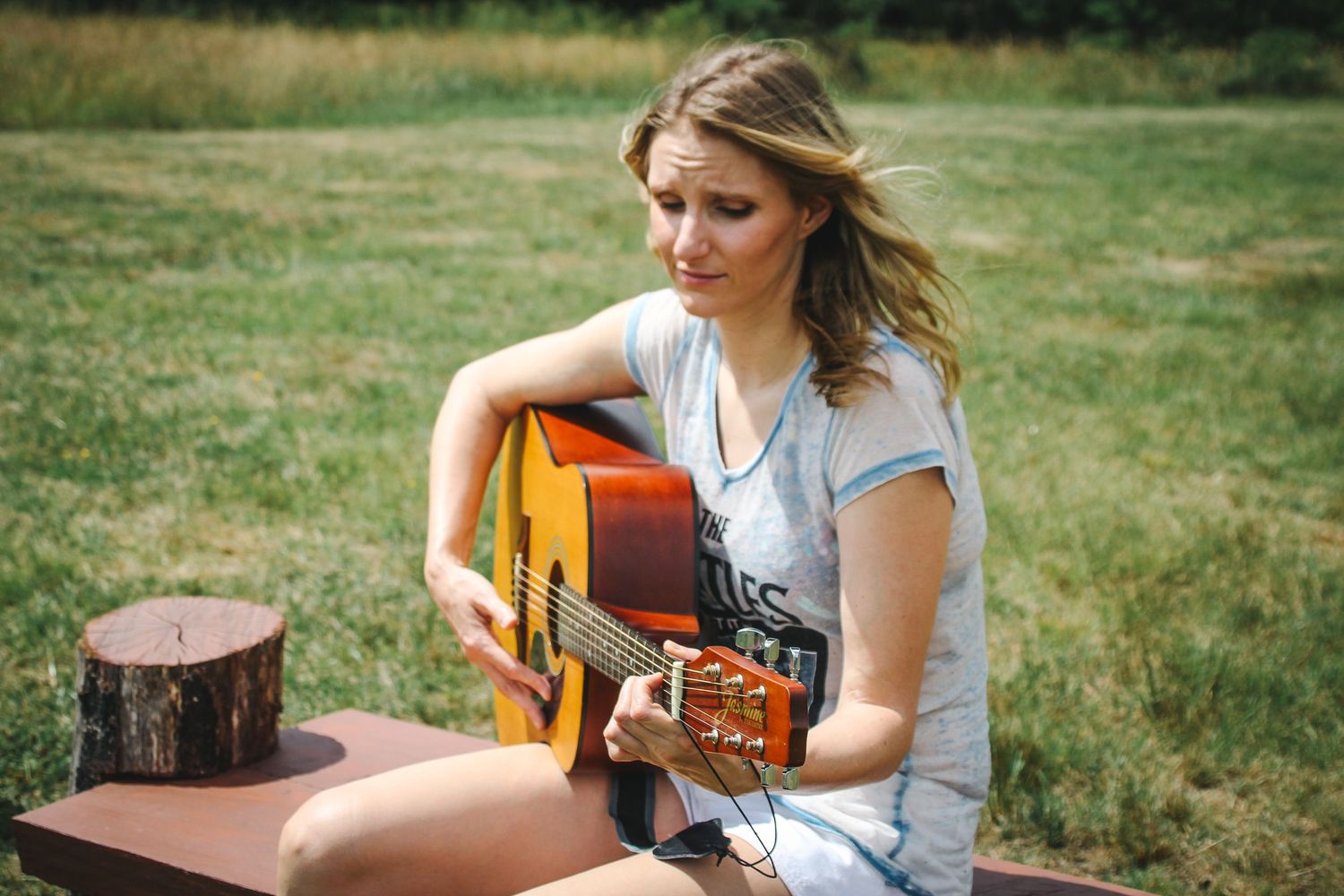 Person playing acoustic guitar outdoors on a sunny day while sitting on wooden stump in grassy field.