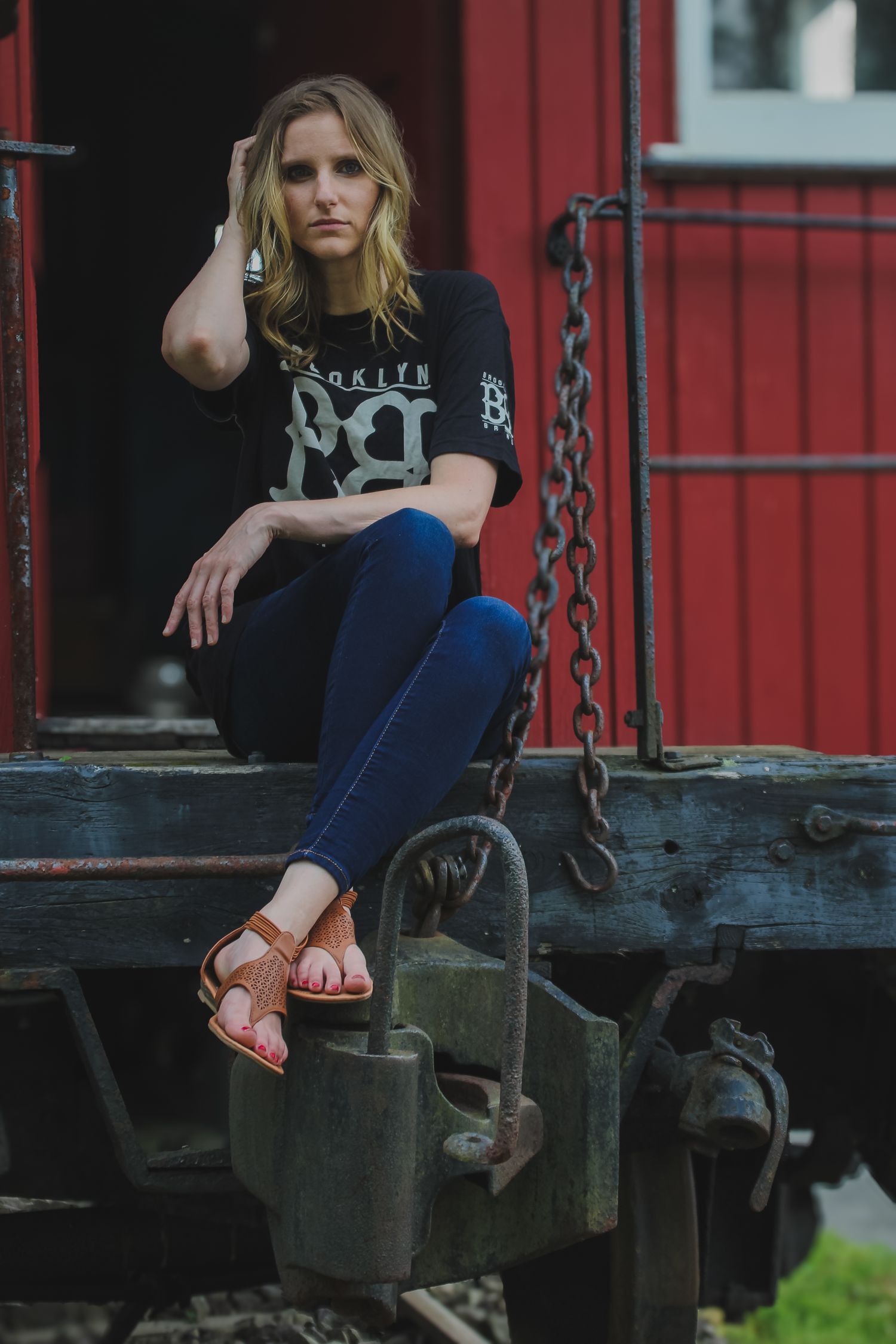 Person in black graphic t-shirt and blue jeans sits casually on red train car wearing brown sandals.