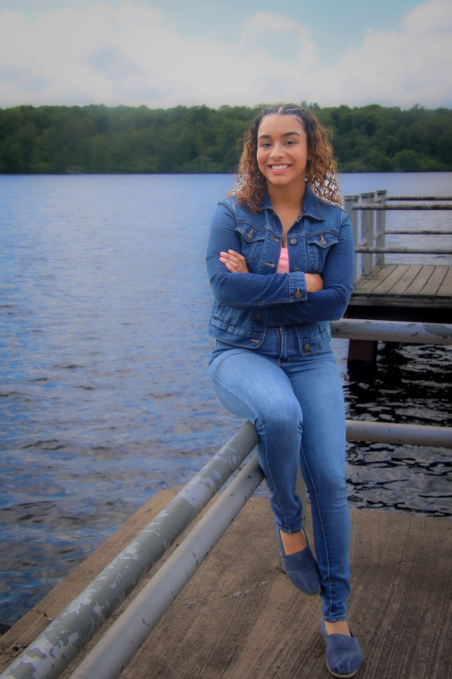 Someone in denim sits on a wooden dock by a serene lake on a sunny day.
